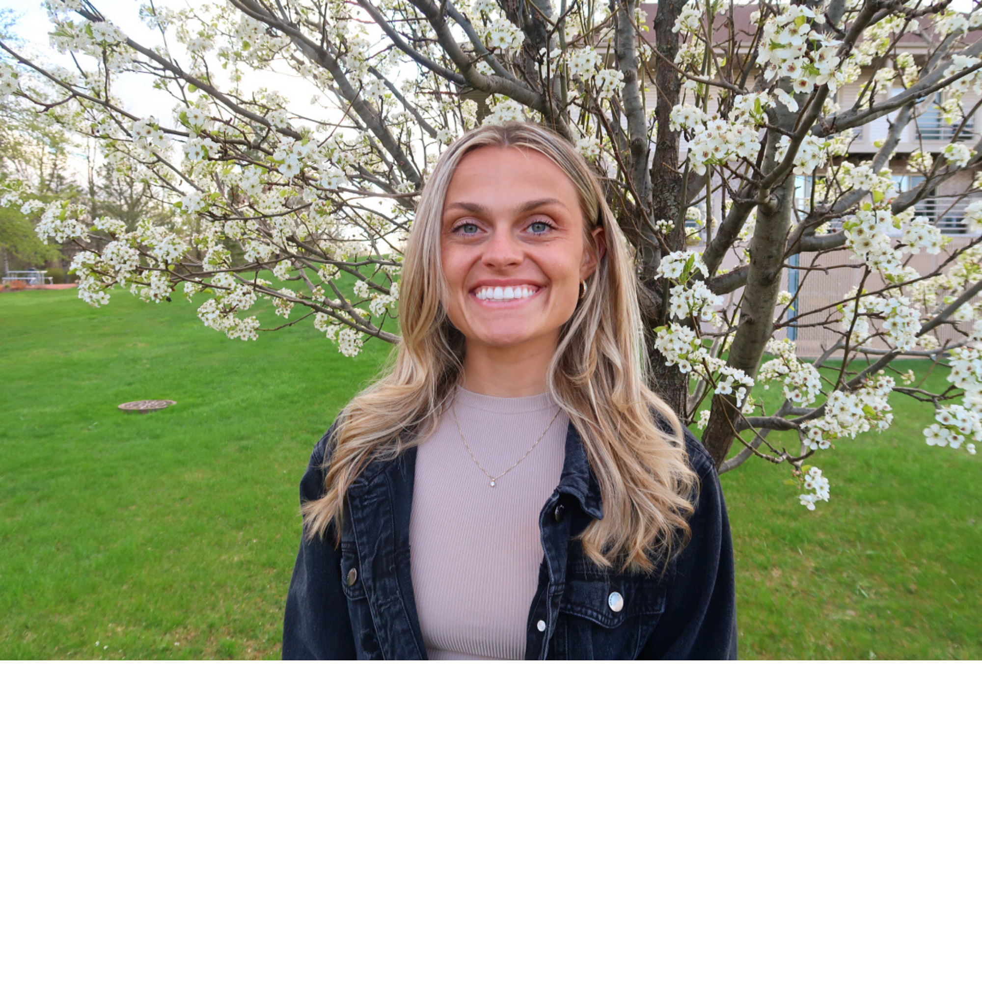 A young woman with long blonde hair and blue eyes standing outdoors in front of a flowering tree with white blossoms, wearing a black denim jacket and a beige top.