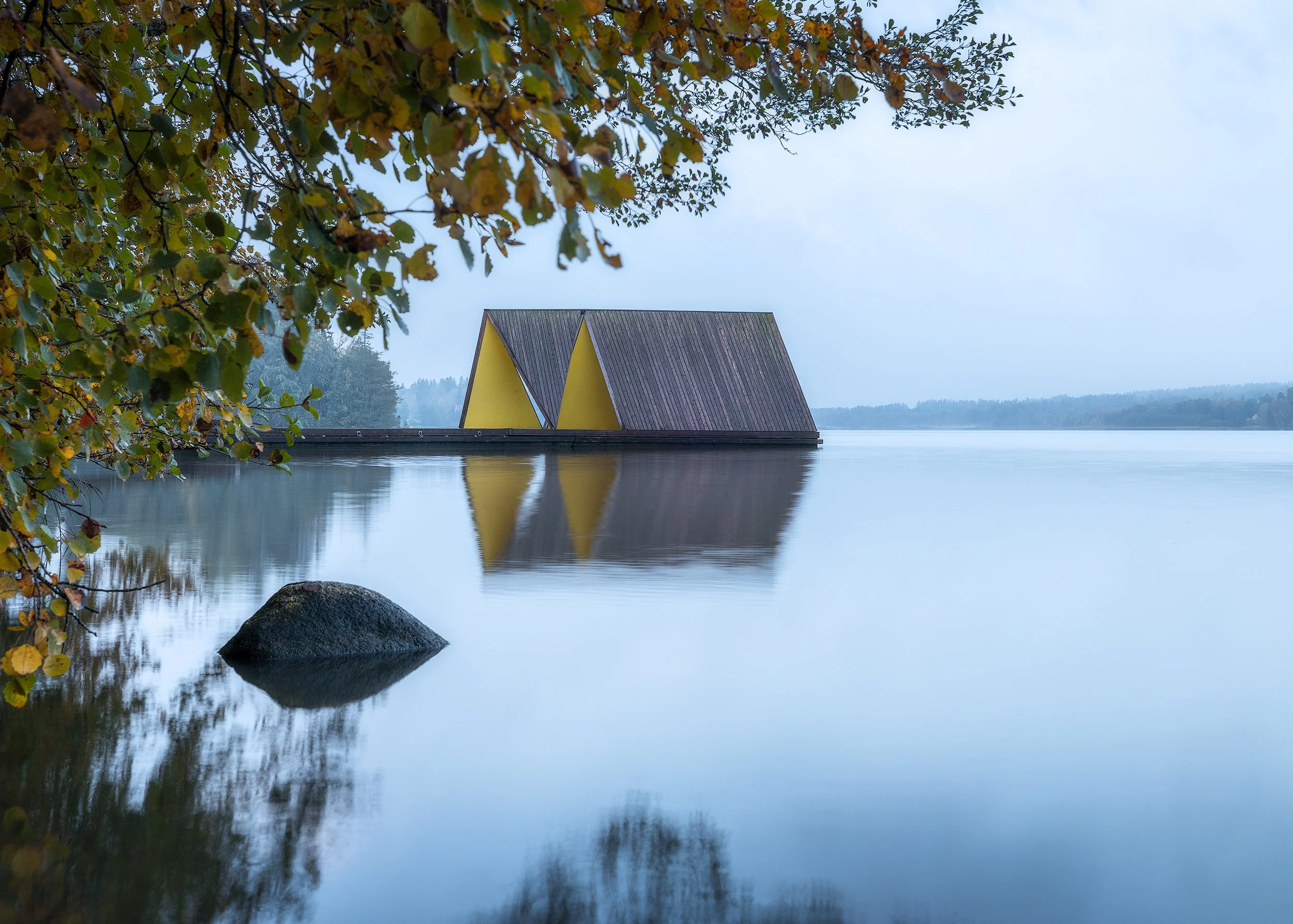 Tuusula floating pavilion reflected in still morning lake, timber and yellow panels