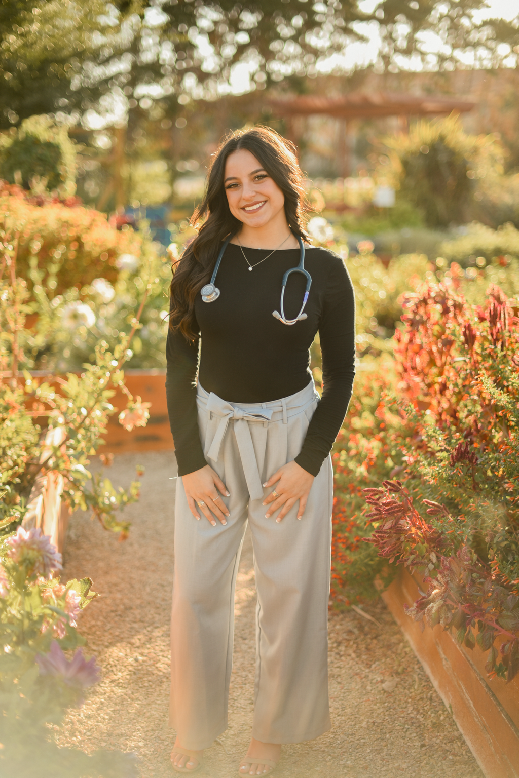 A woman with long dark hair smiling at a garden center during golden hour, wearing a black top, beige wide-leg pants with a bow, and a stethoscope around her neck.