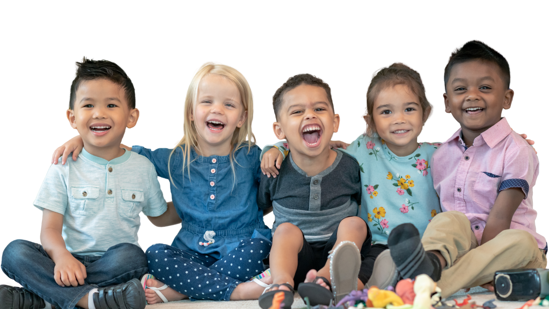 A group of six diverse happy children sitting close together, with arms around each other, smiling and laughing, dressed casually, with toys in front of them.