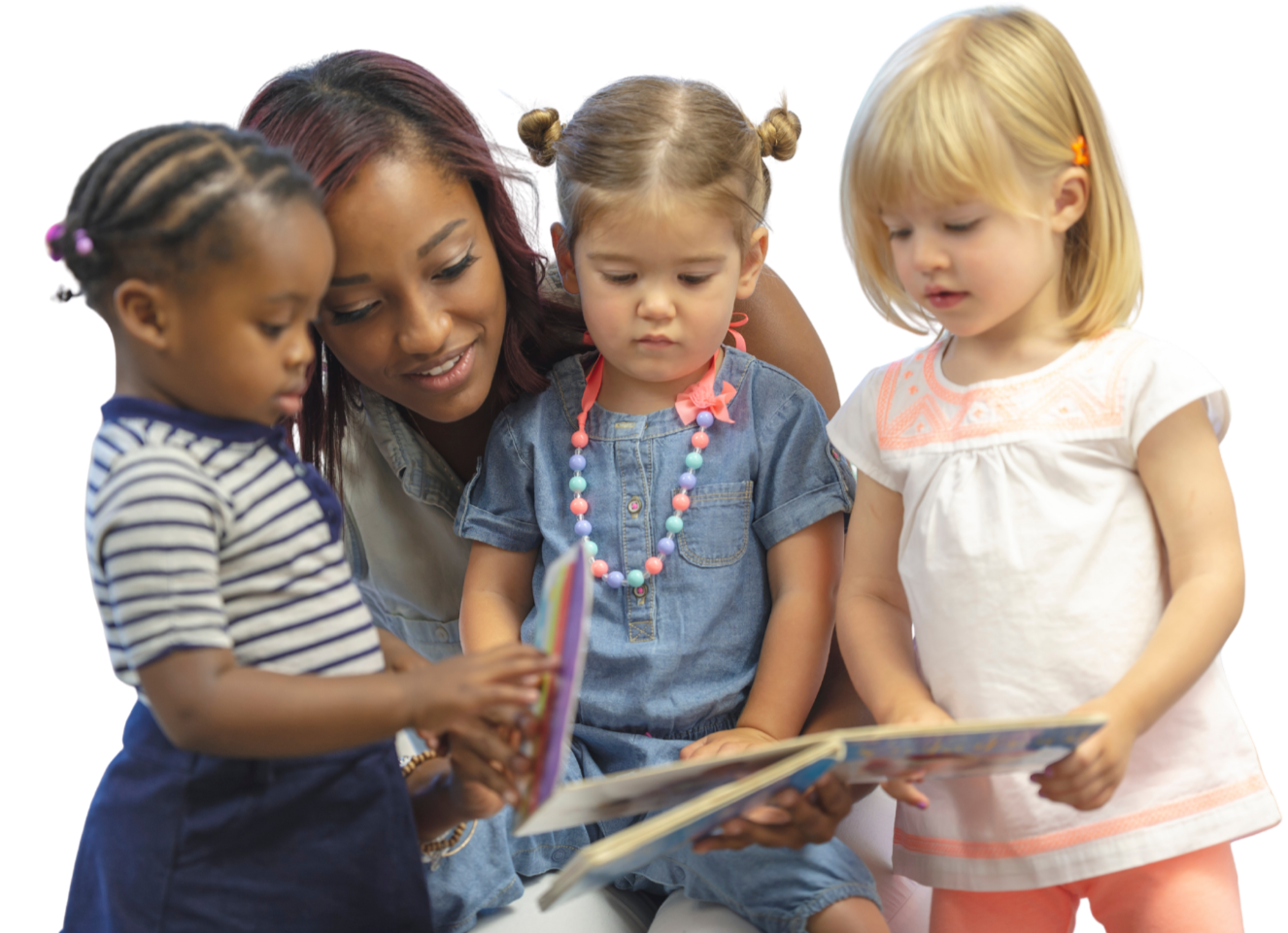 An adult woman reading a children's book with three young girls, one in a striped shirt, another in a denim dress with a colorful necklace, and the third in a white dress, all looking at the book together.