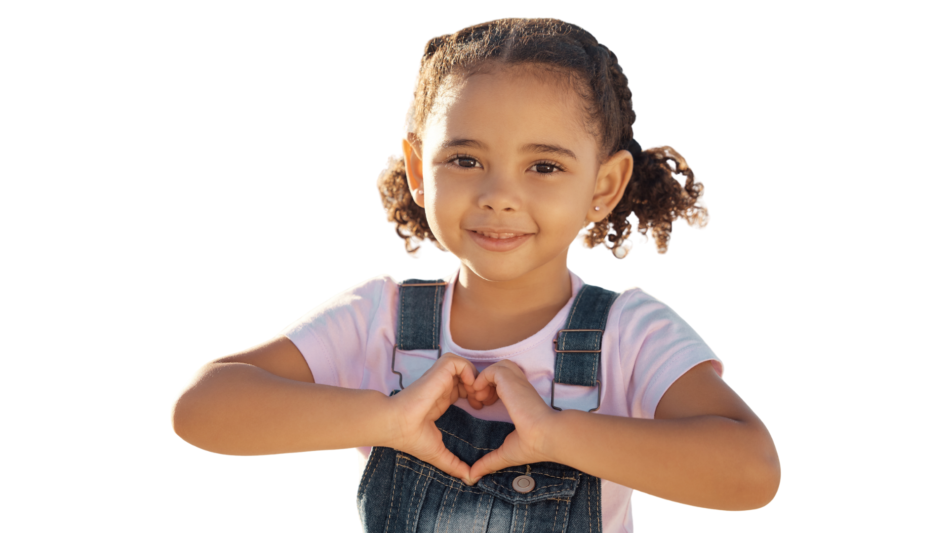 A young girl with curly hair in pigtails smiling, wearing a pink shirt and denim overalls, making a heart shape with her hands.