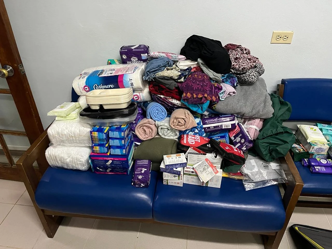 A hospital waiting room bench filled with various supplies including folded blankets, packs of adult diapers, small packages of medical gloves, and other healthcare items.