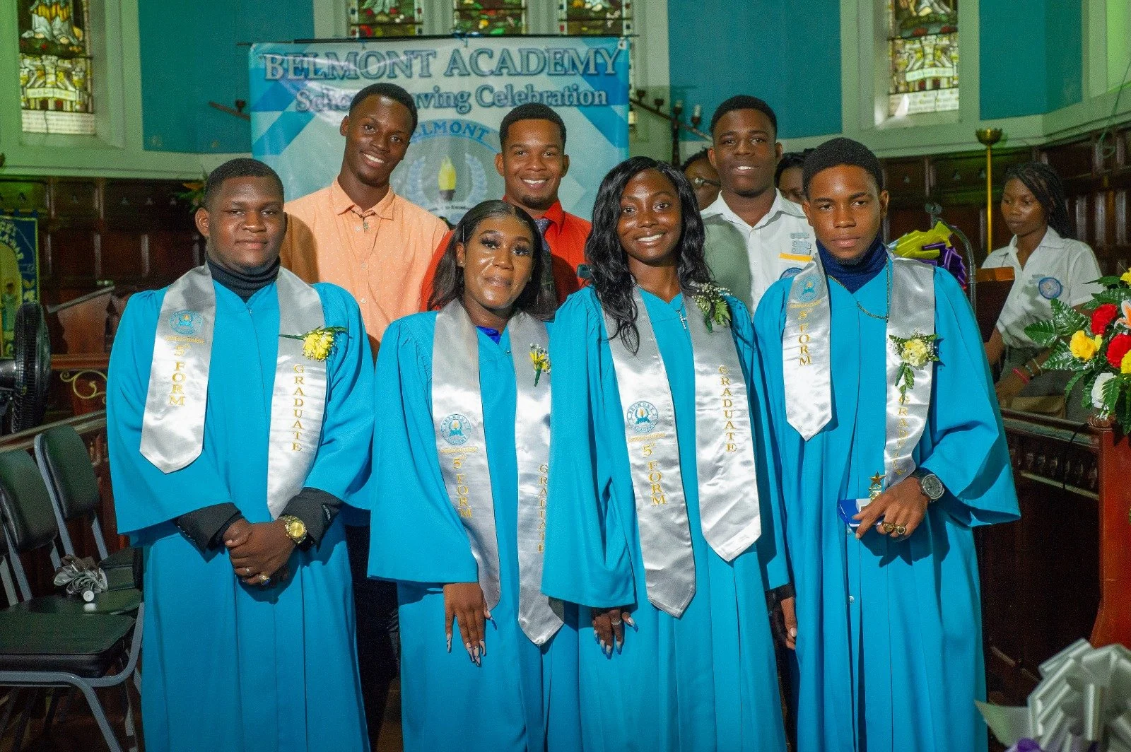 Group of graduates wearing blue caps and gowns with silver stoles, celebrating a graduation ceremony inside a church with stained glass windows.
