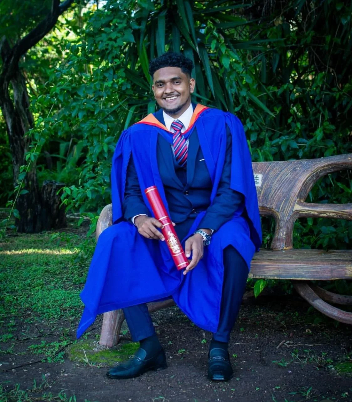 A young man in a navy blue graduation gown and cap, with an orange and blue hood, sitting on a wooden bench outdoors in front of green foliage. He is holding a red diploma tube and is smiling at the camera.