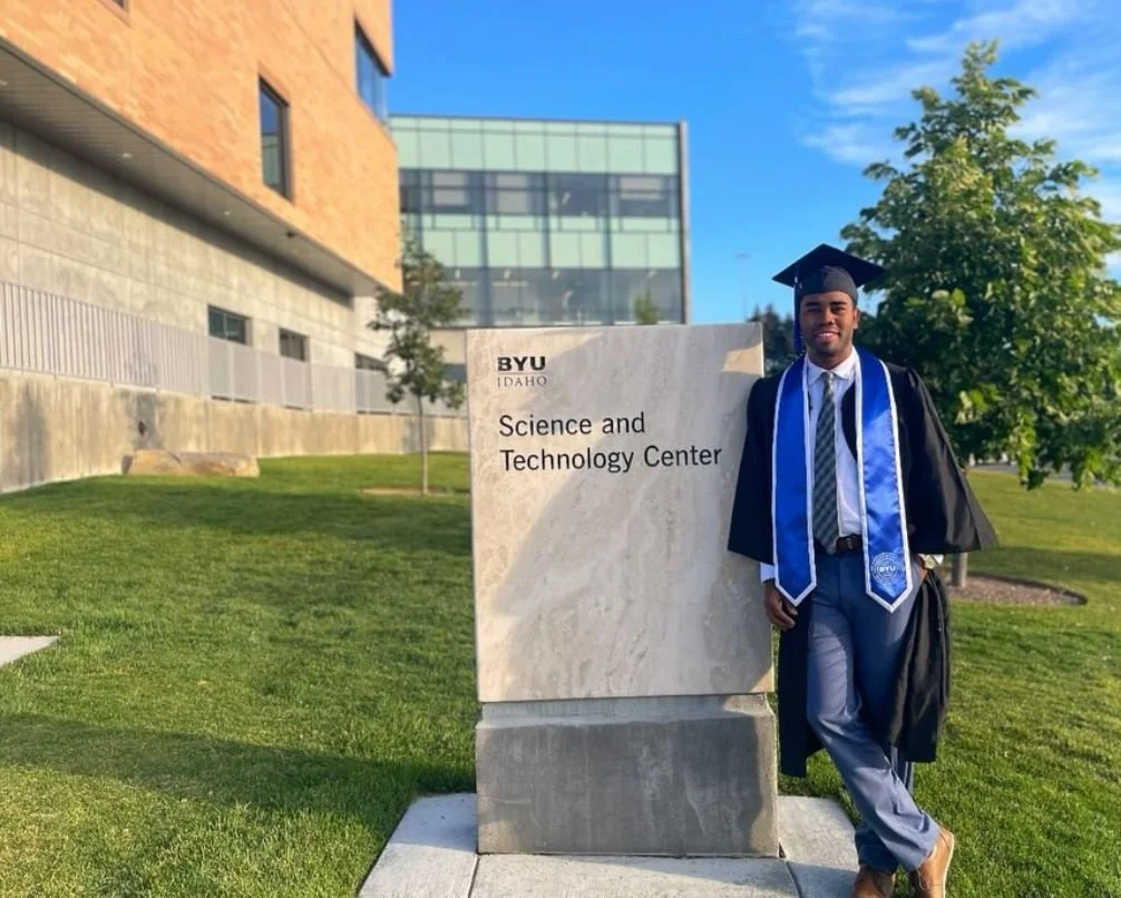 Graduate in cap and gown standing next to a sign that reads 'BYU Idaho Science and Technology Center' outside on a school campus with green grass and trees.
