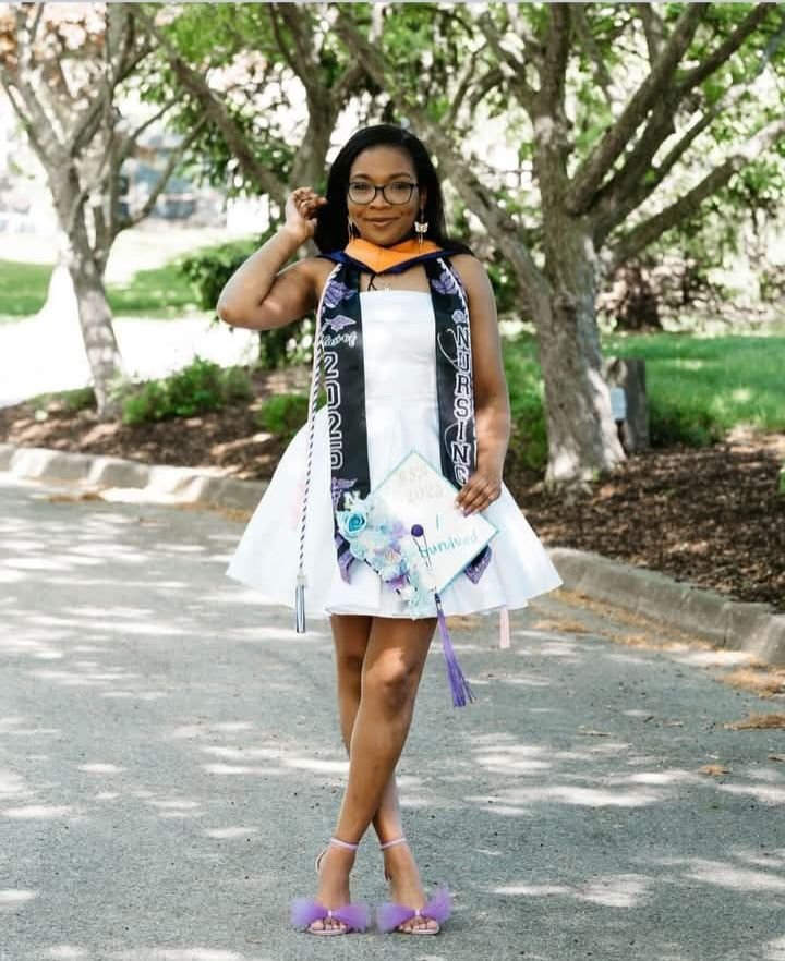 Young woman in a white dress and graduation stole, holding a graduation cap, standing outdoors on a paved path with trees in the background.
