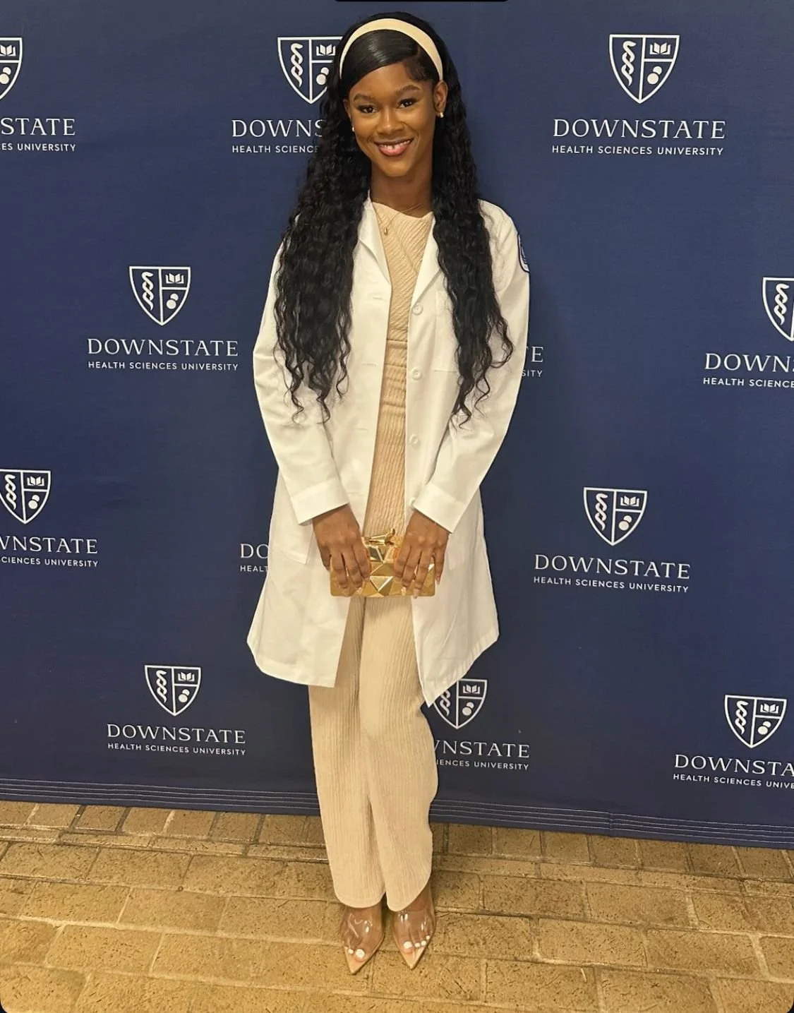 A woman in a beige outfit with a white coat, curly hair, and a headband holding a golden clutch, standing in front of a blue backdrop with the Downstate Health Sciences University logo.