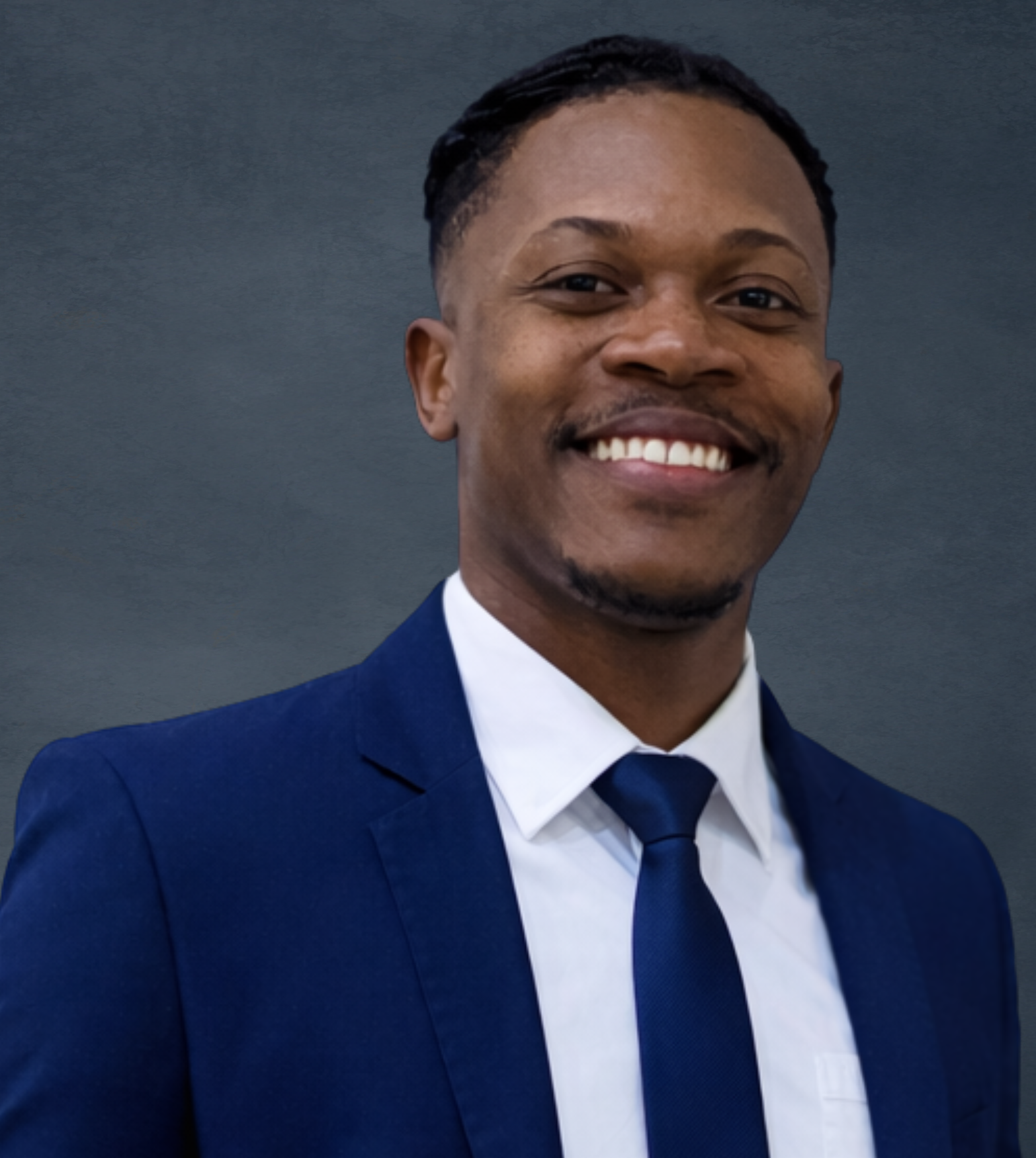 A smiling man in a dark blue suit, white shirt, and matching blue tie against a gray background.
