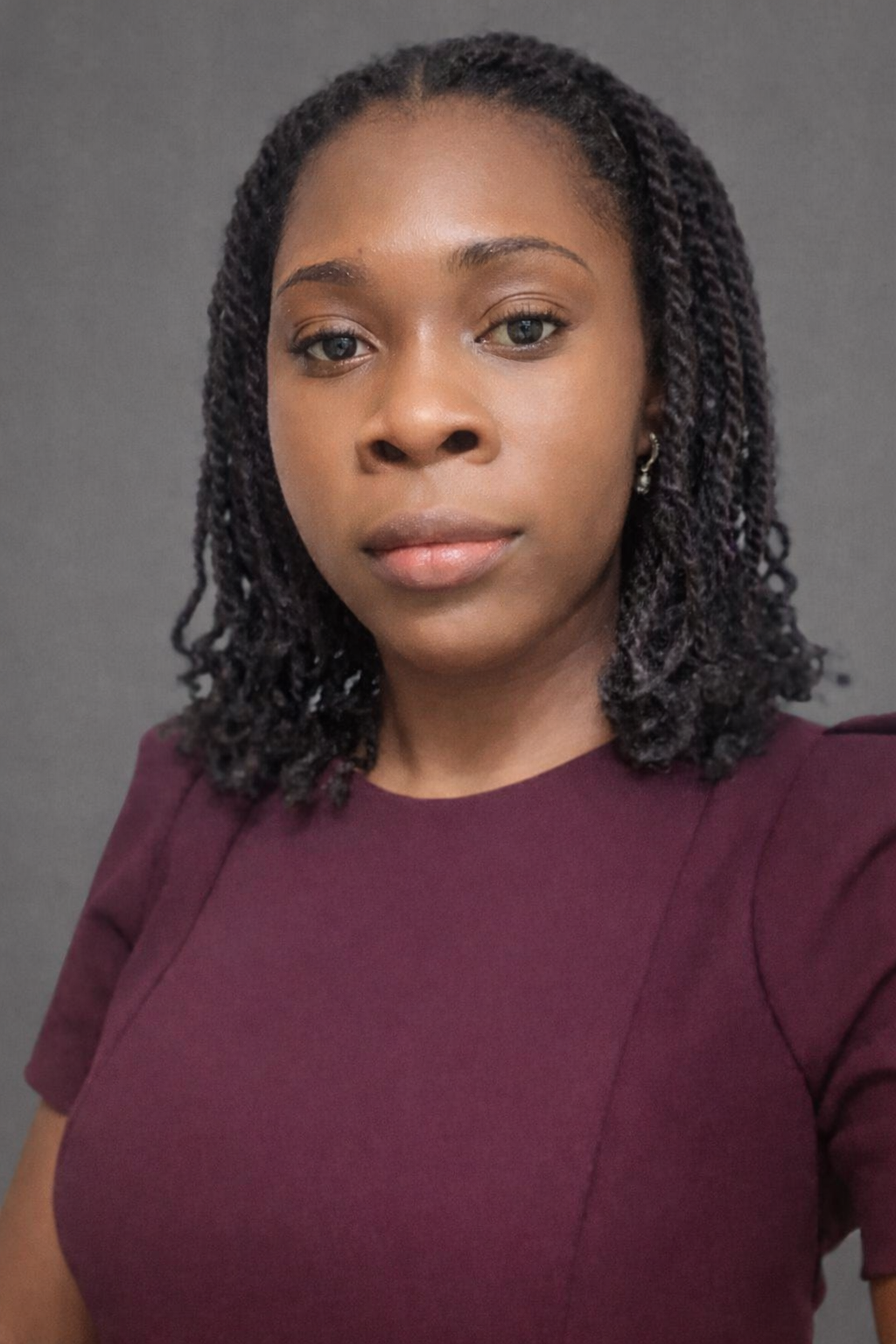 A woman with dark skin, shoulder-length curly hair, wearing a maroon top, looking directly at the camera against a gray background.