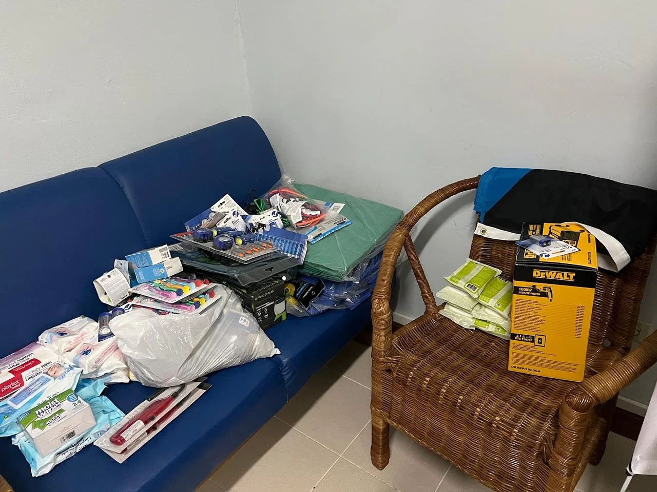 Piles of various supplies, including cleaning products, tools, and hardware, stored on a blue bench and a wicker chair in a small room.