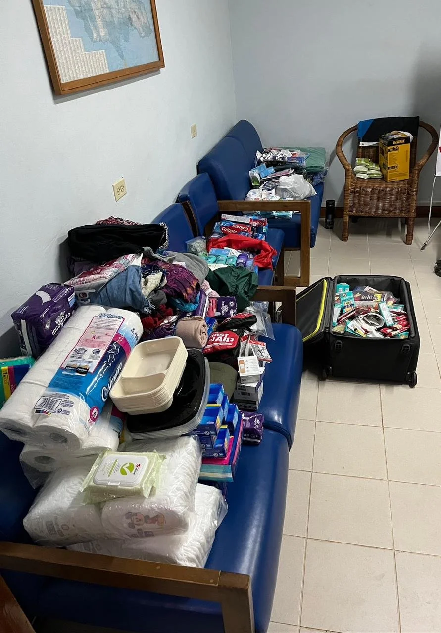 A waiting room filled with various hygiene and household supplies, including toilet paper, disposable plates, toothpaste, and personal care items, spread across blue chairs and a black suitcase.