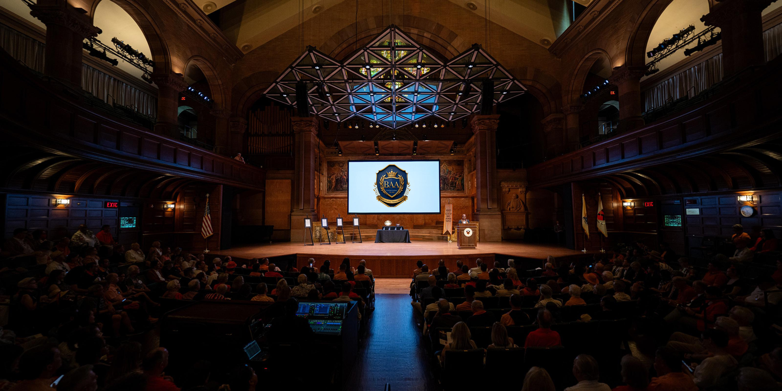 A large auditorium filled with people attending a formal event or ceremony, with a stage displaying a screen with a shield and the letters 'BAA'. The room has high ceilings and ornate wood paneling, with flags and artwork visible.