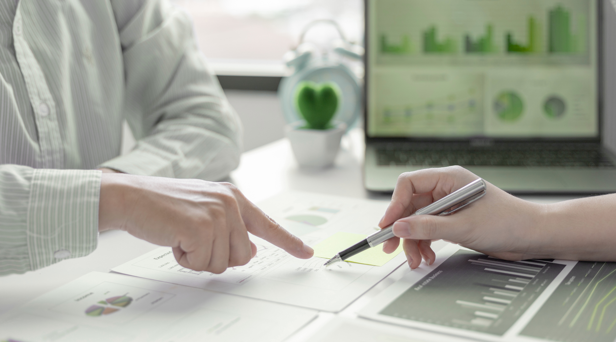 Two people pointing at financial documents and charts on a desk, with a laptop displaying graphs and a decorative green heart on a plant in the background.