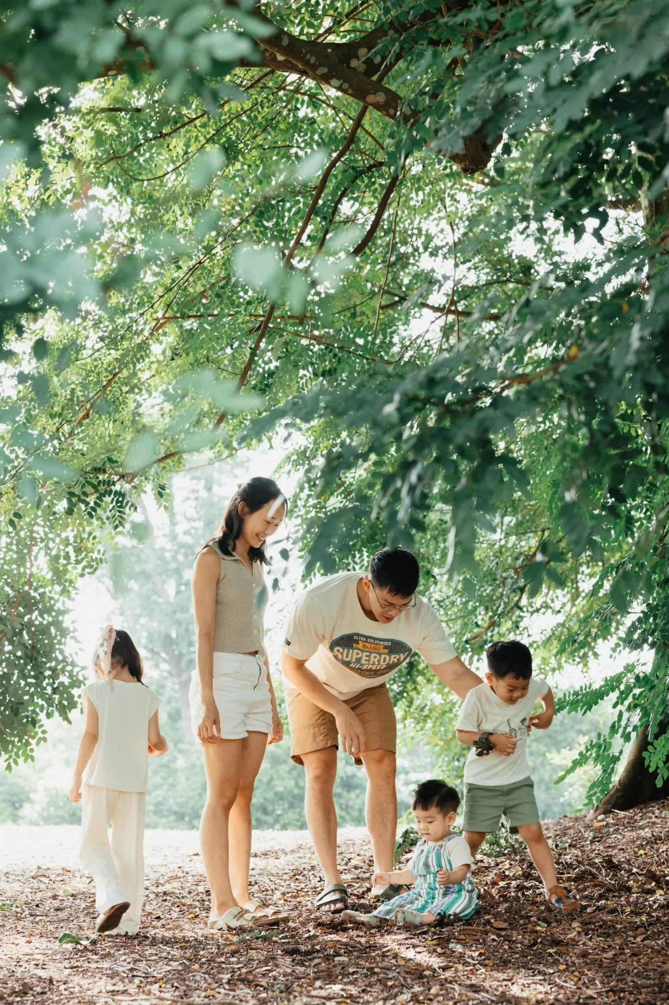 A family of five, including two children, gather outdoors under a leafy tree during daytime. The family members are smiling and involved in picking up fallen leaves or acorns from the ground.