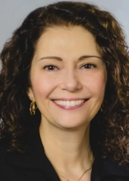 A woman with medium-length curly hair, light skin, and gold earrings, smiling at the camera.