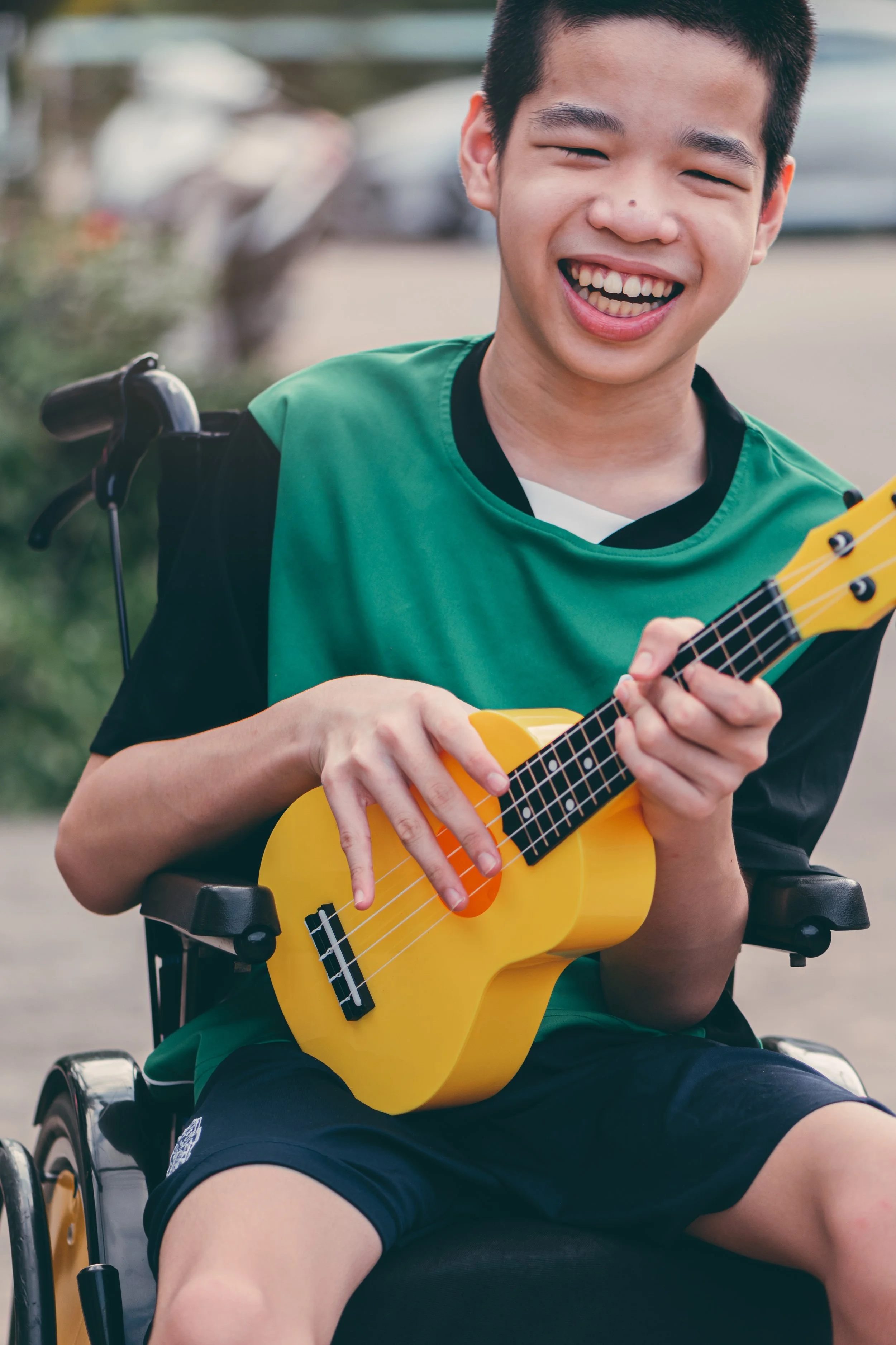 A smiling boy in a wheelchair playing a yellow ukulele outdoors.