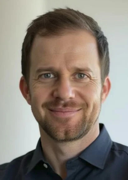 A man with short brown hair, light skin, and a slight beard smiling, wearing a dark collared shirt, standing indoors with a light-colored wall behind him.