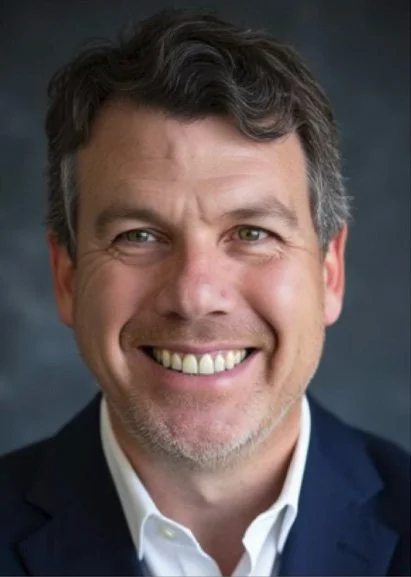 Close-up of a middle-aged man smiling, with dark hair and wearing a dark blazer and white shirt.
