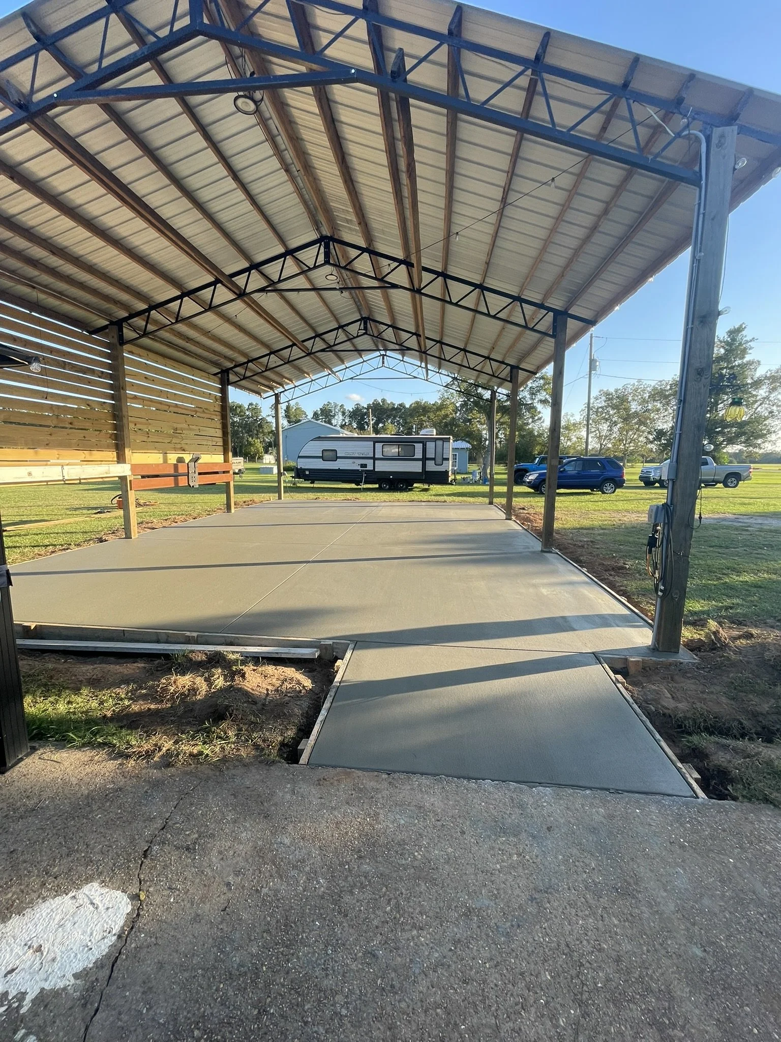 A covered parking area with a concrete floor, surrounded by grass, with a camper trailer and vehicles visible in the background.