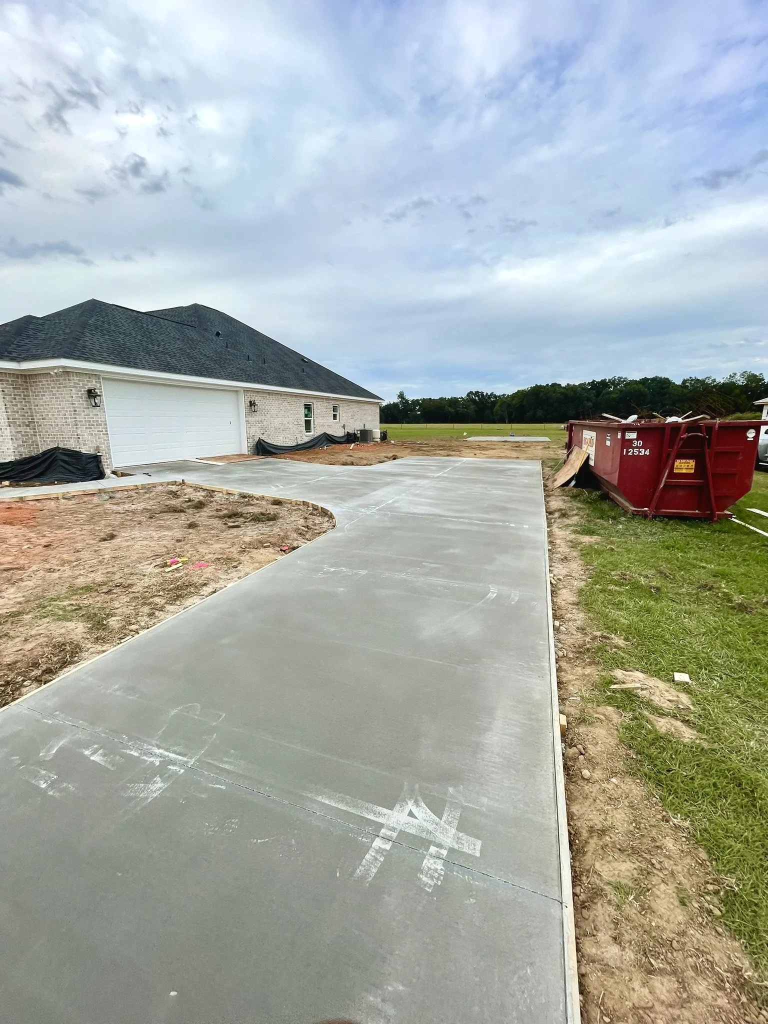 Newly poured concrete sidewalk leading to a house with a garage, construction materials, and a red dumpster on the right side, under a cloudy sky.