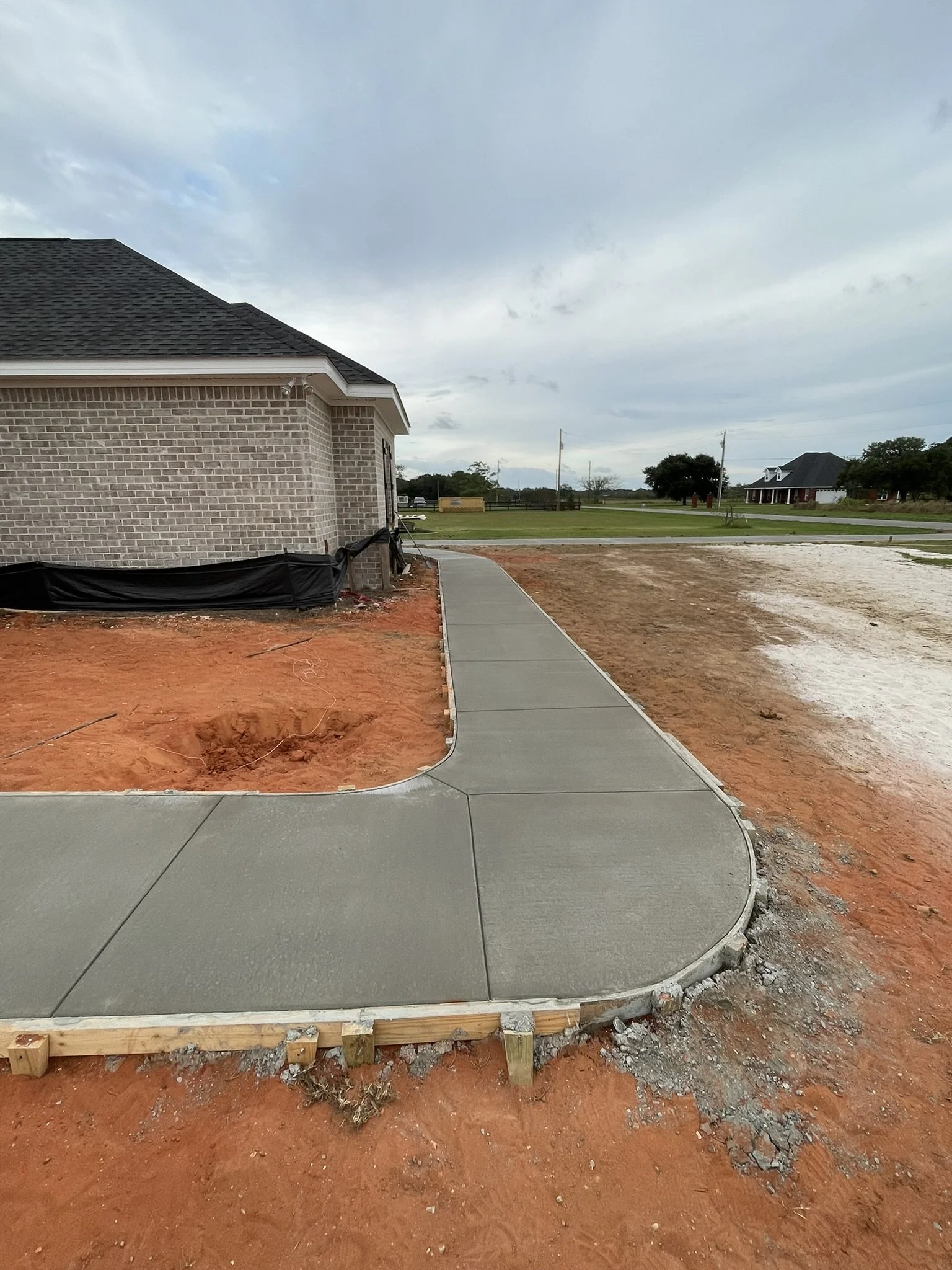 Finished concrete sidewalk with curb in front of a house under construction, with red dirt and building materials around.