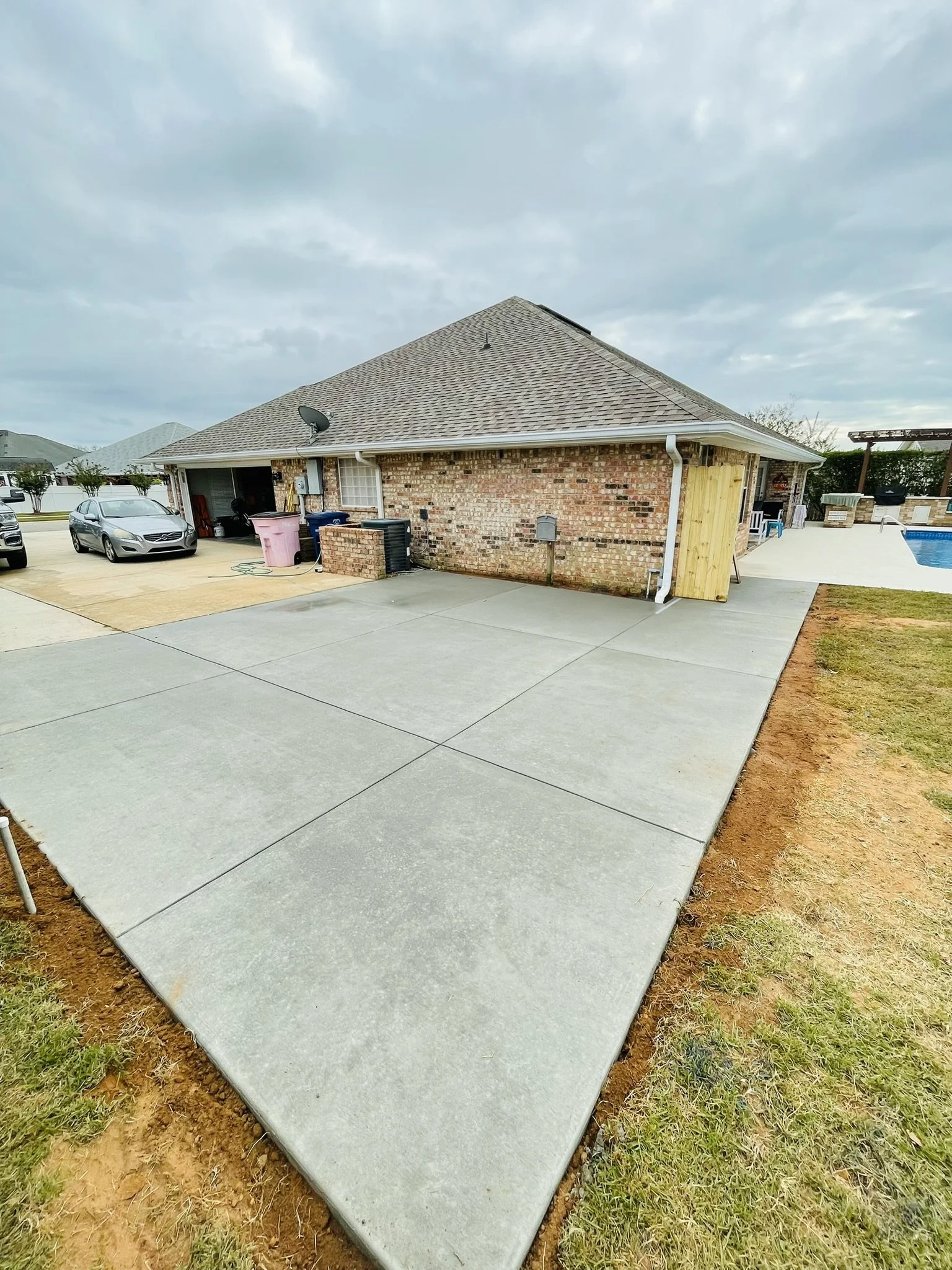 A backyard patio area with newly poured concrete, with a brick house and neighboring houses visible in the background. The sky is cloudy.