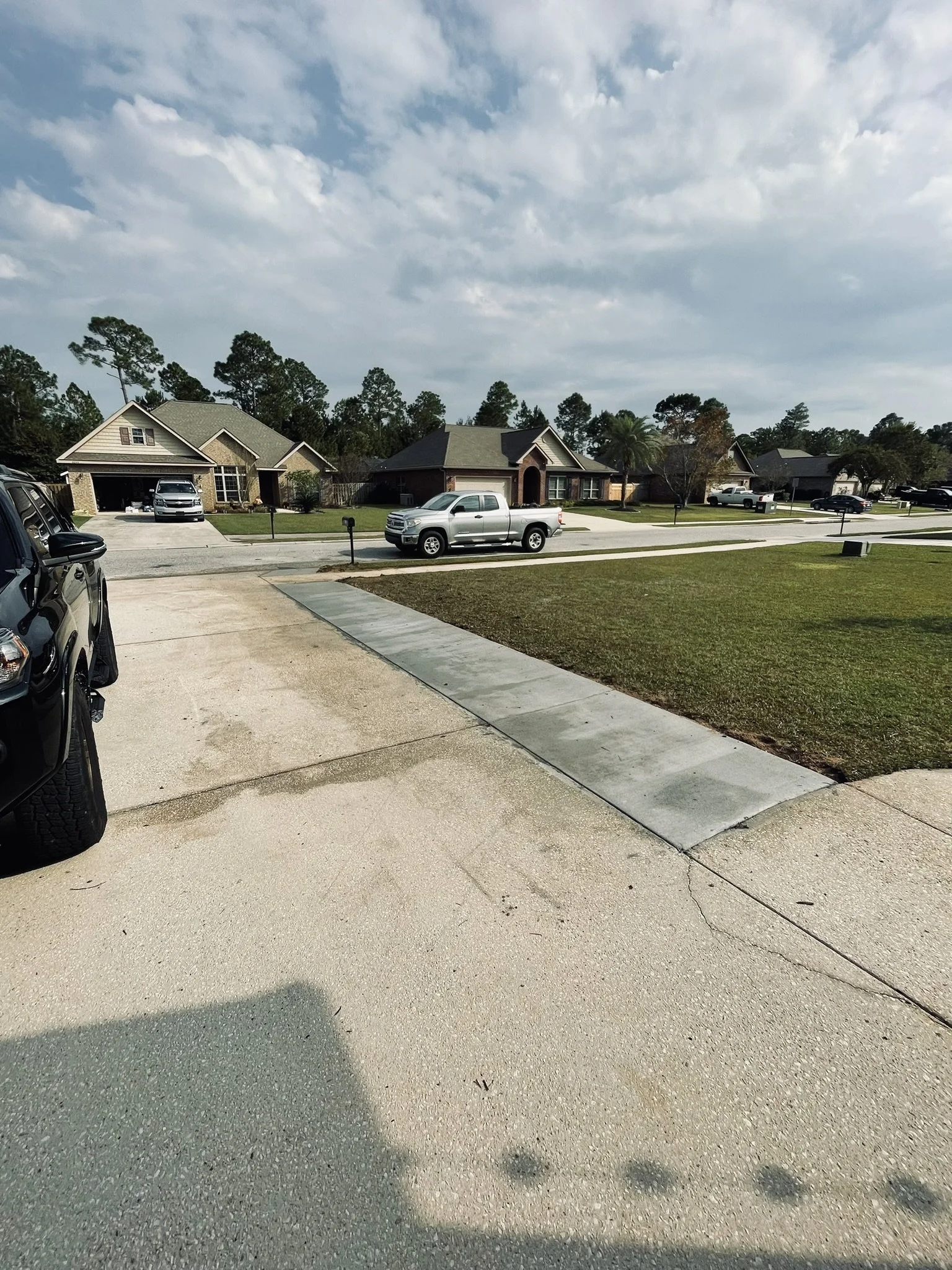A suburban neighborhood street with parked cars, green lawns, and single-family houses under a partly cloudy sky.