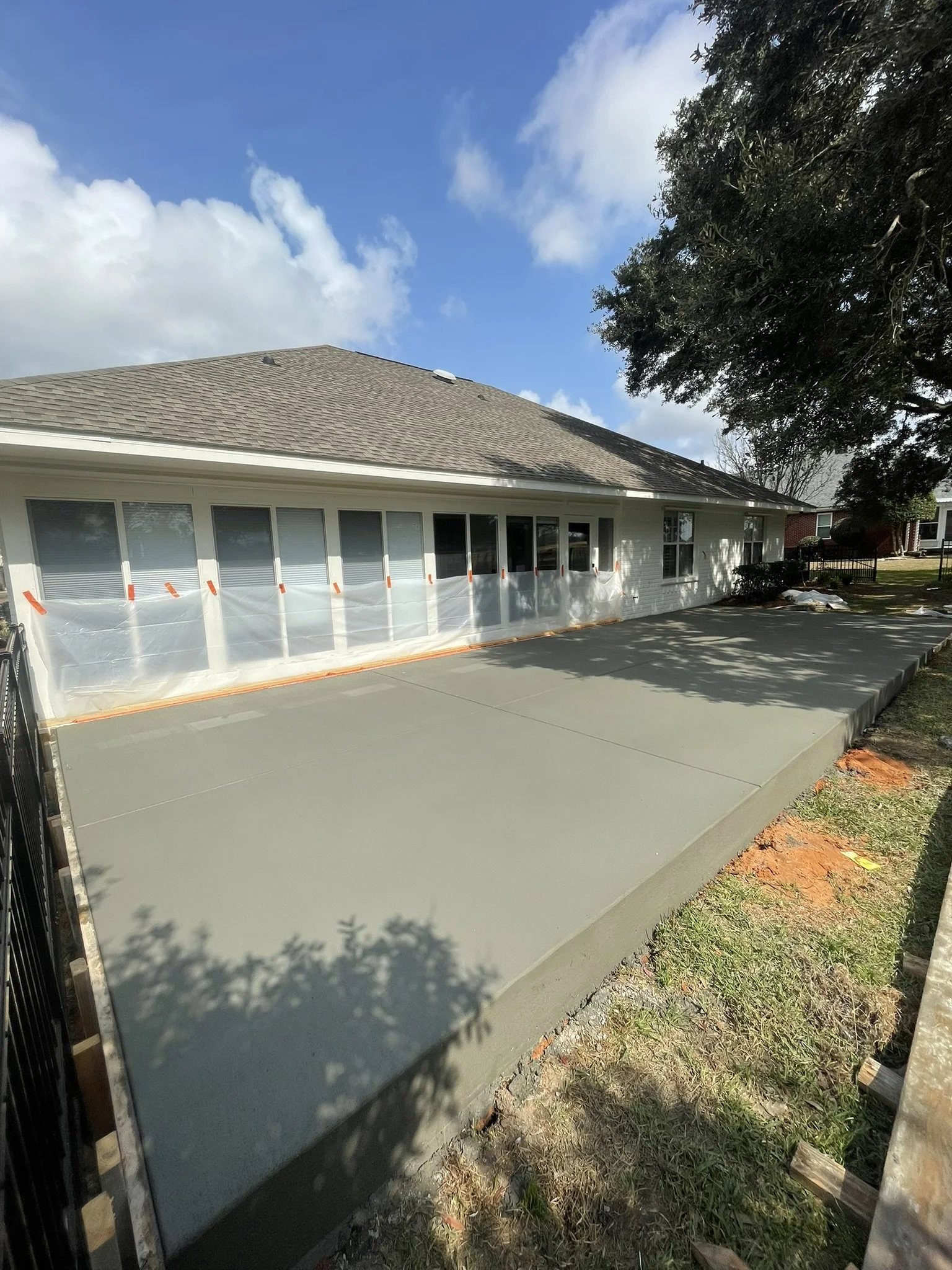 Freshly poured concrete slab on backyard patio with a house in the background, plastic sheeting and orange tape covering windows