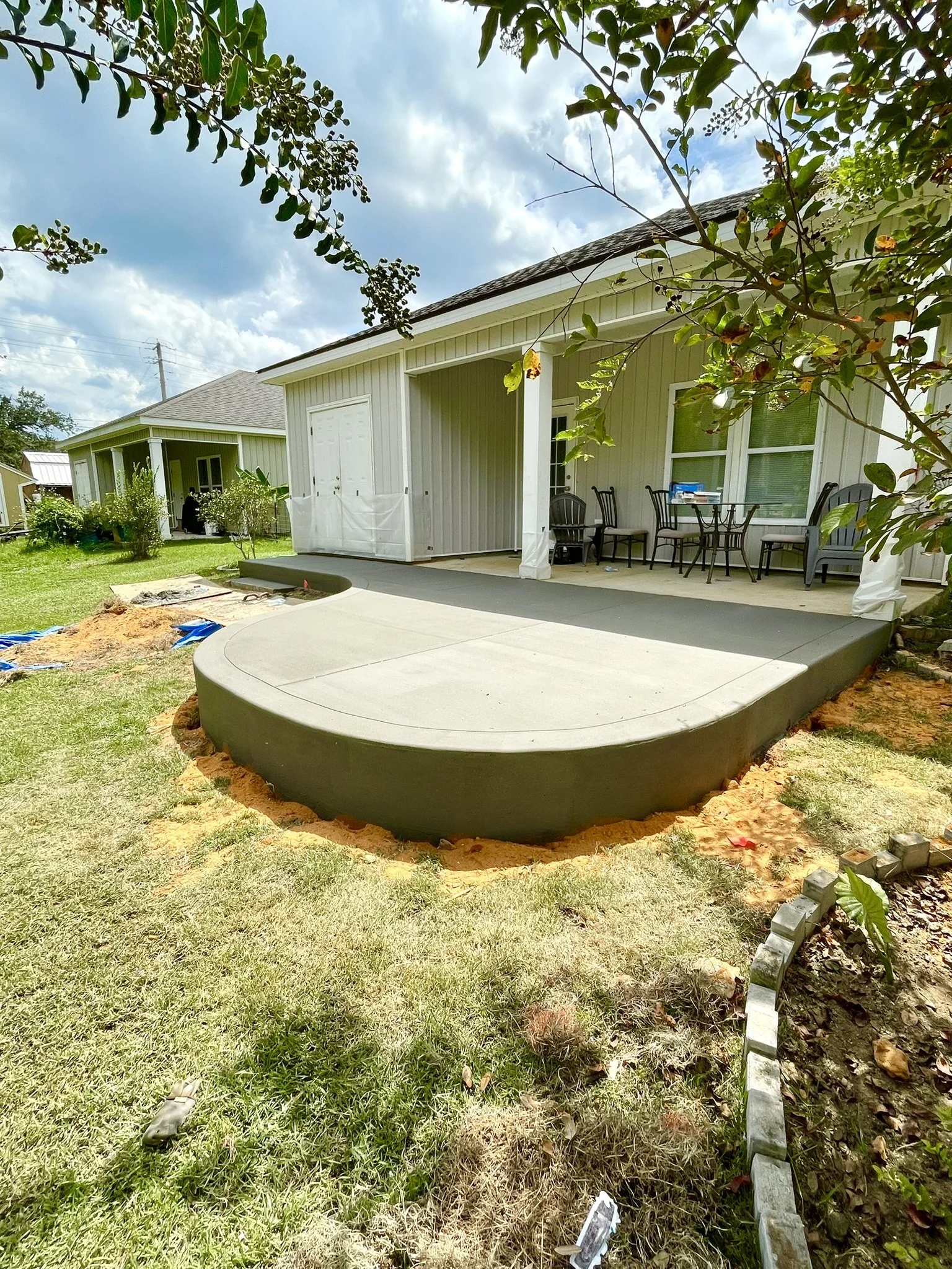 Freshly poured concrete patio with a rounded edge in a backyard, attached to a house with a porch and outdoor furniture, surrounded by grass and trees.