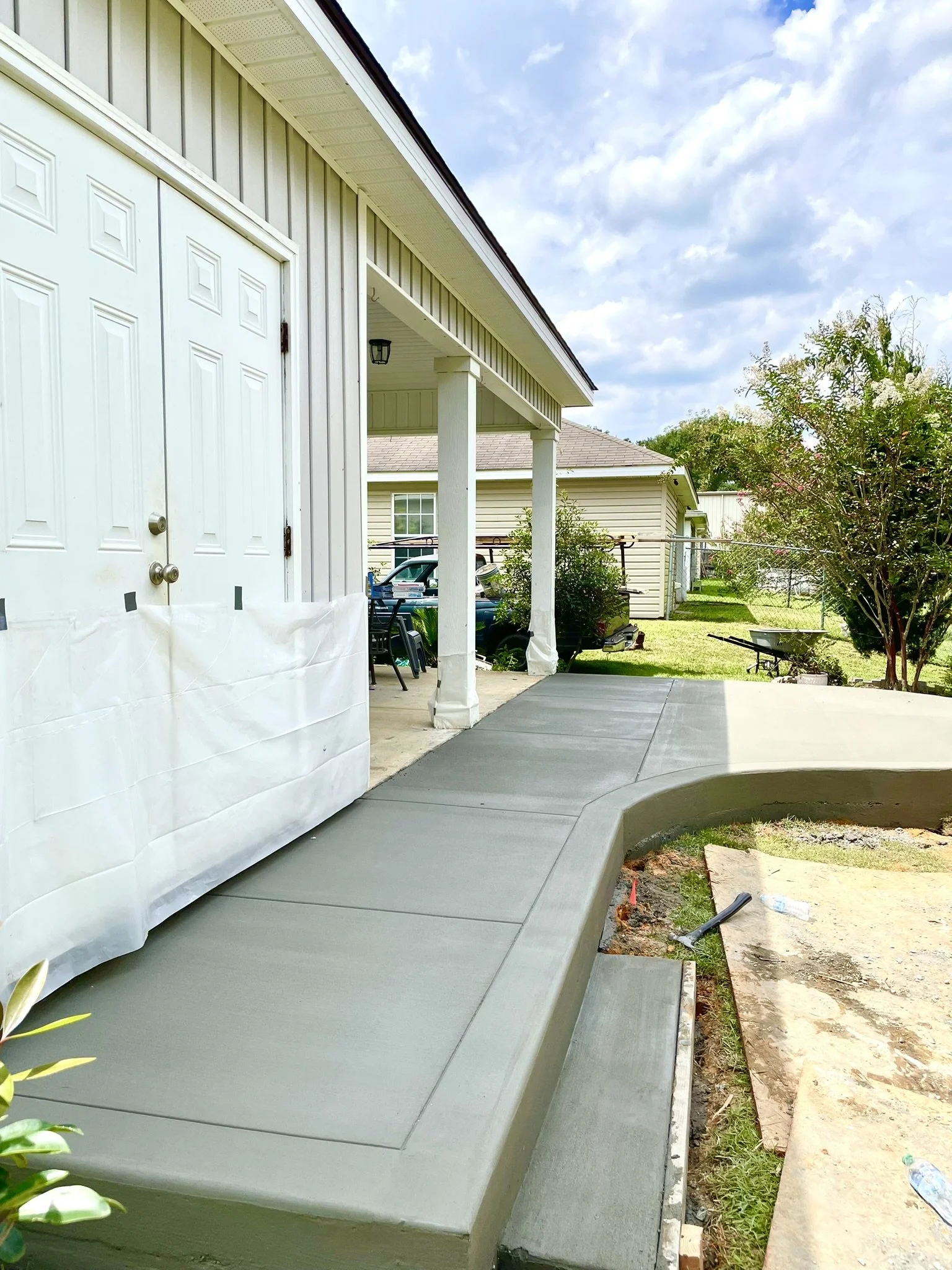 Newly poured concrete patio and steps alongside a house with a white garage door, patio furniture, and a backyard with trees and grass.
