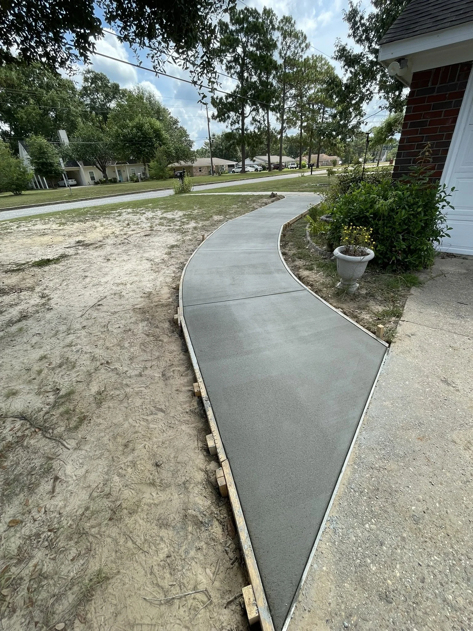Newly poured concrete sidewalk with a curved shape, installed beside a brick house with a white garage door, in a residential neighborhood with trees and houses in the background.