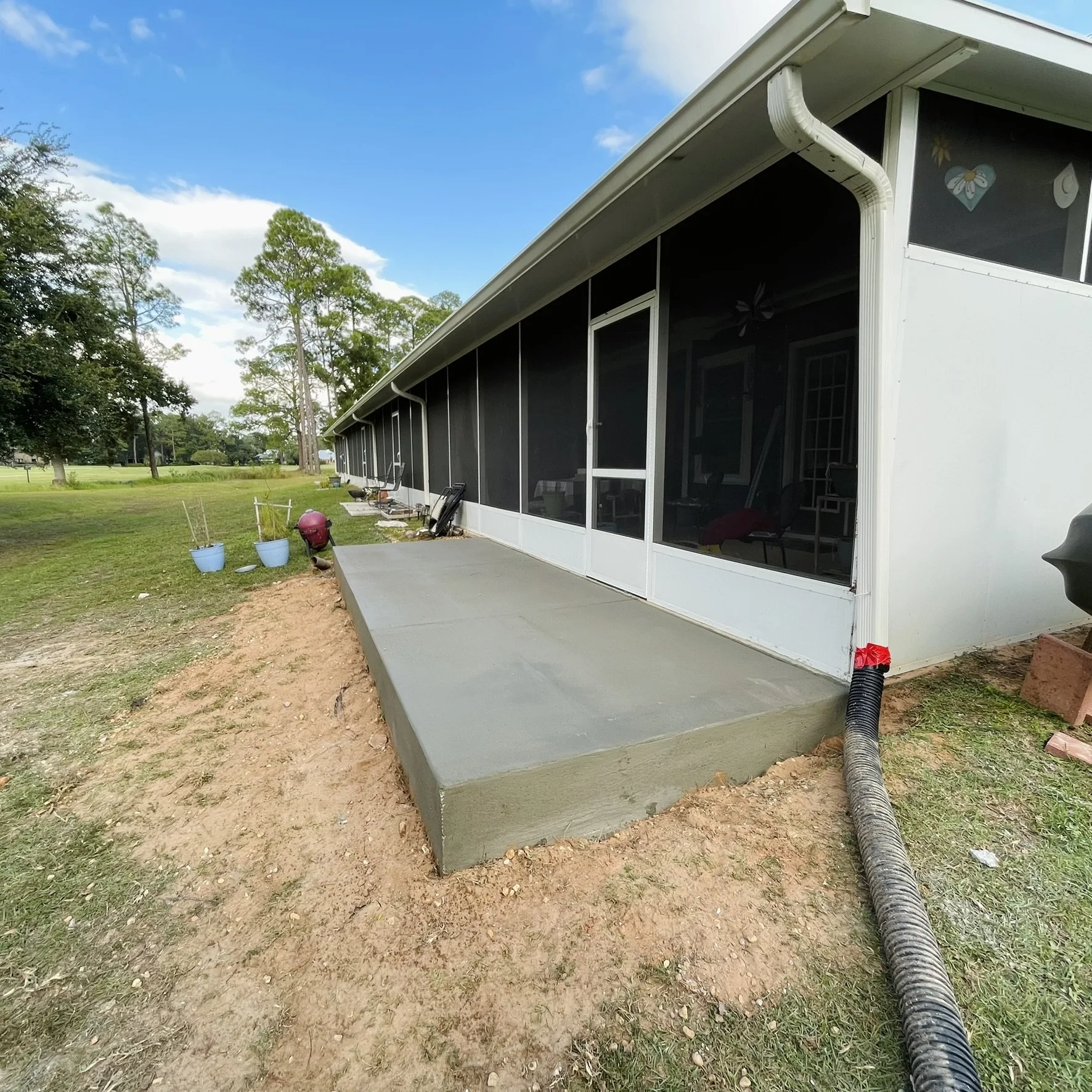 A newly poured concrete patio next to a white house with a screened-in porch, on a grassy yard with trees in the background, under a partly cloudy sky.