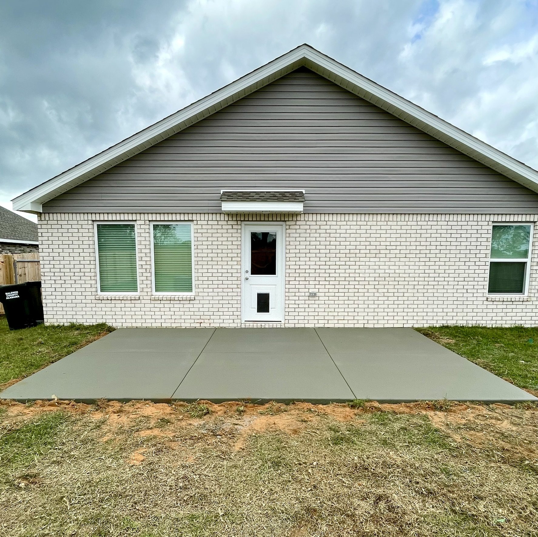 Back of a house with a concrete patio and two windows, brick and siding exterior, door with pet door, cloudy sky.