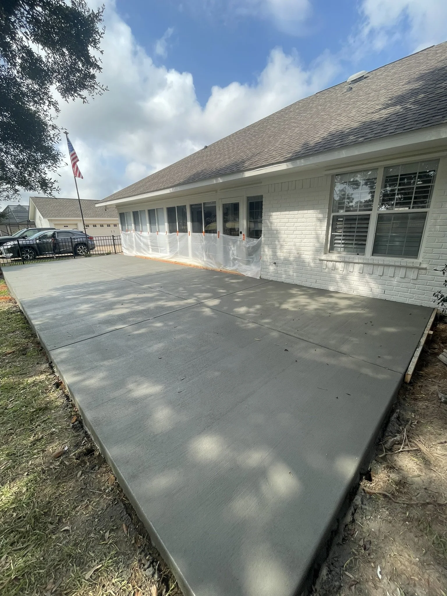 Newly poured concrete patio in the backyard of a house with white brick walls, large windows, and a shingled roof. A flagpole with an American flag is visible in the background. The patio is surrounded by a small patch of grass and a dirt area.