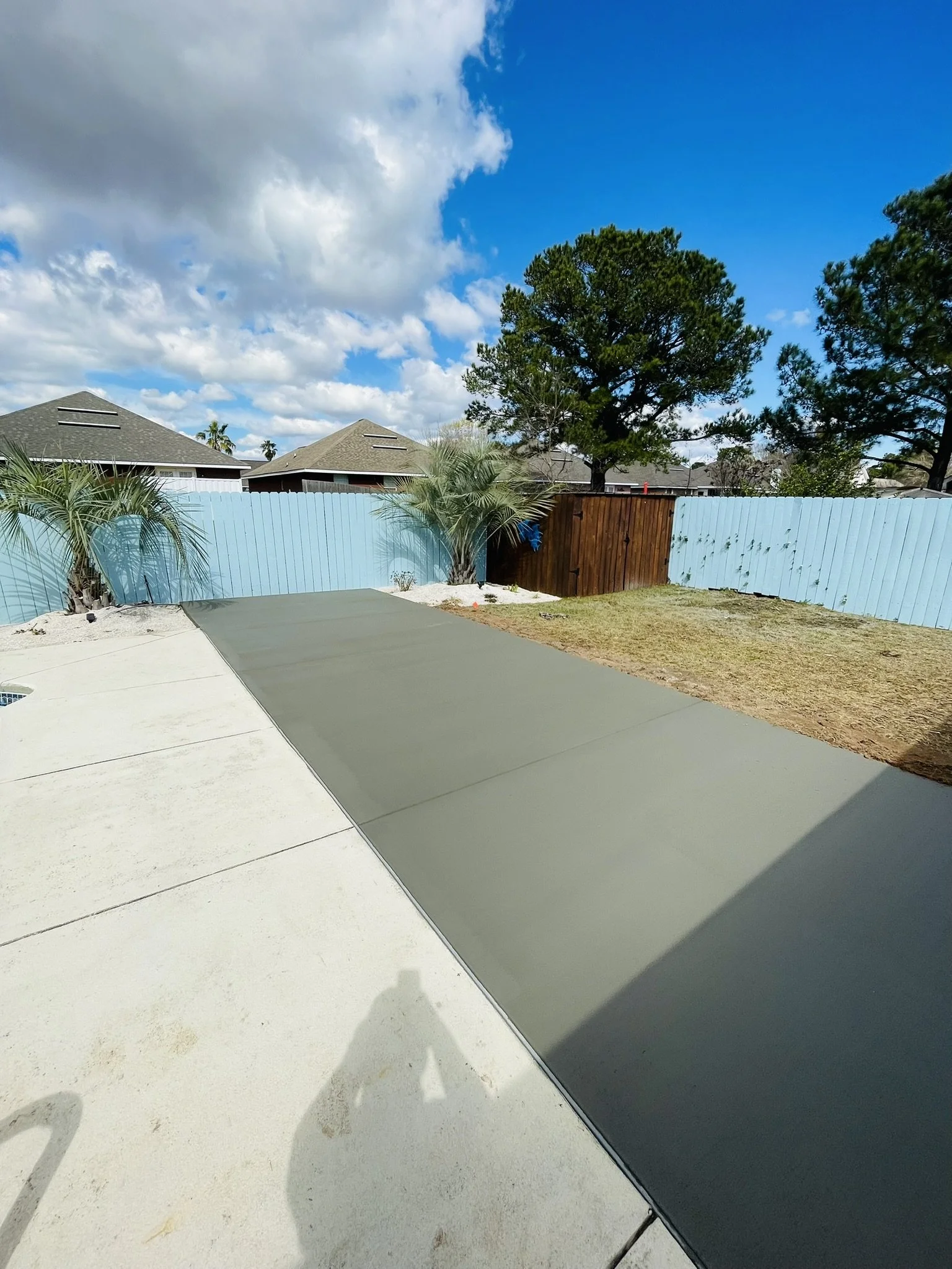 A backyard with a concrete patio, a blue fence, some palm trees, and a section of new asphalt pavement under the sky with clouds.