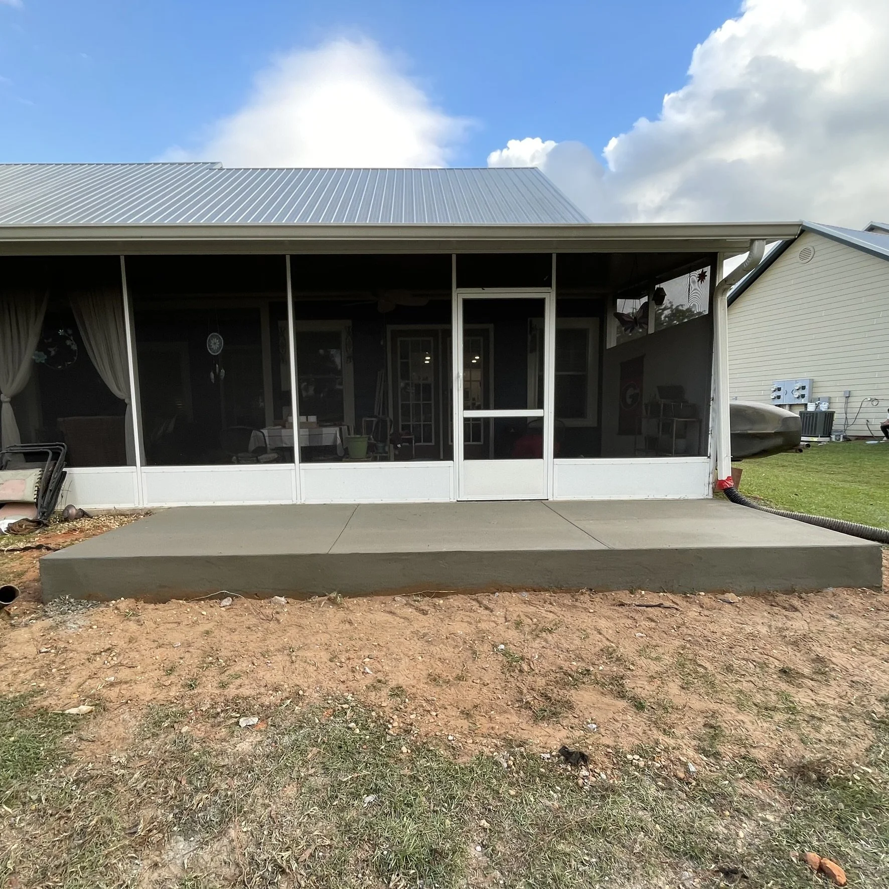 Backyard patio with newly poured concrete slab in front of a screened-in porch with glass doors, outdoor furniture, and decorations. Grass and soil in the foreground, neighboring house with a green lawn and outdoor equipment in the background.