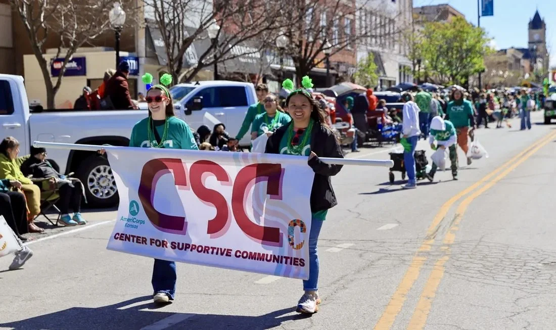 People participating in a parade, holding a banner that reads 'Center for Supportive Communities,' with some wearing green accessories and shamrock hats, on a city street during a sunny day.