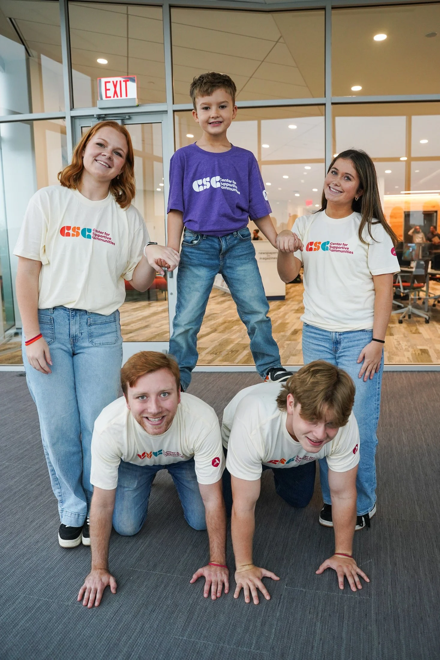 Group of young people in a gym or community center making a human pyramid. Three people are on the ground on all fours, supporting the person above. The person on top is a young boy standing and holding hands with two women, one on each side, who are also supporting him.
