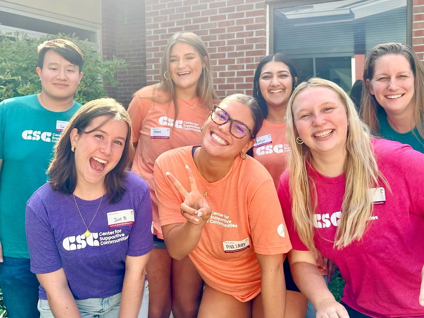 A group of seven people standing outside, group photo, smiling, wearing colorful t-shirts with a 'Center for Supportive Communities' logo, some showing their name tags, sunny day.