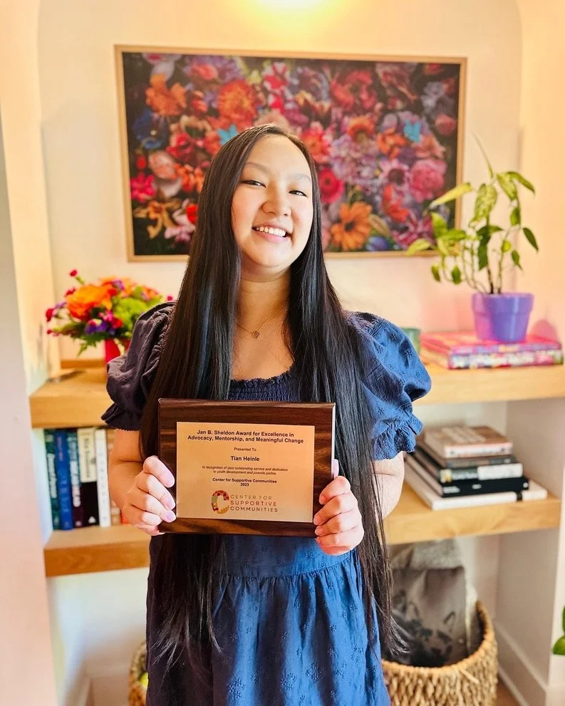 Young woman with long dark hair smiling and holding a plaque, standing in a room with a bookshelf, colorful flowers, and a framed floral artwork on the wall.