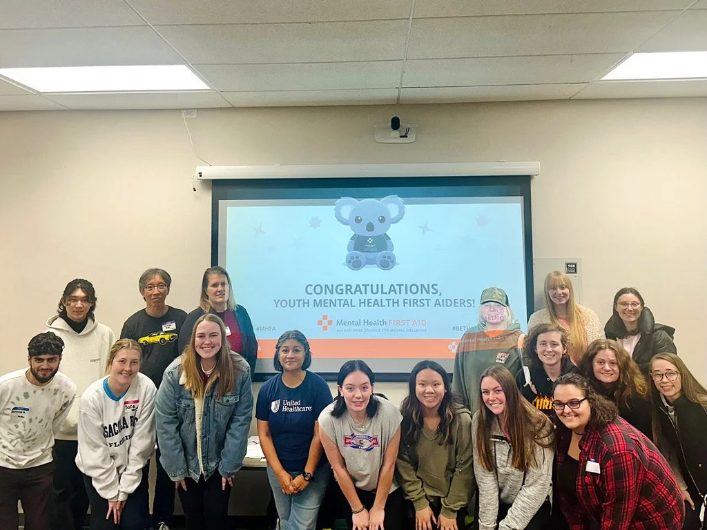 Group of young people and adults posing in a classroom in front of a projection screen displaying a congratulatory message for Youth Mental Health First Aiders, with a cartoon koala bear illustration.