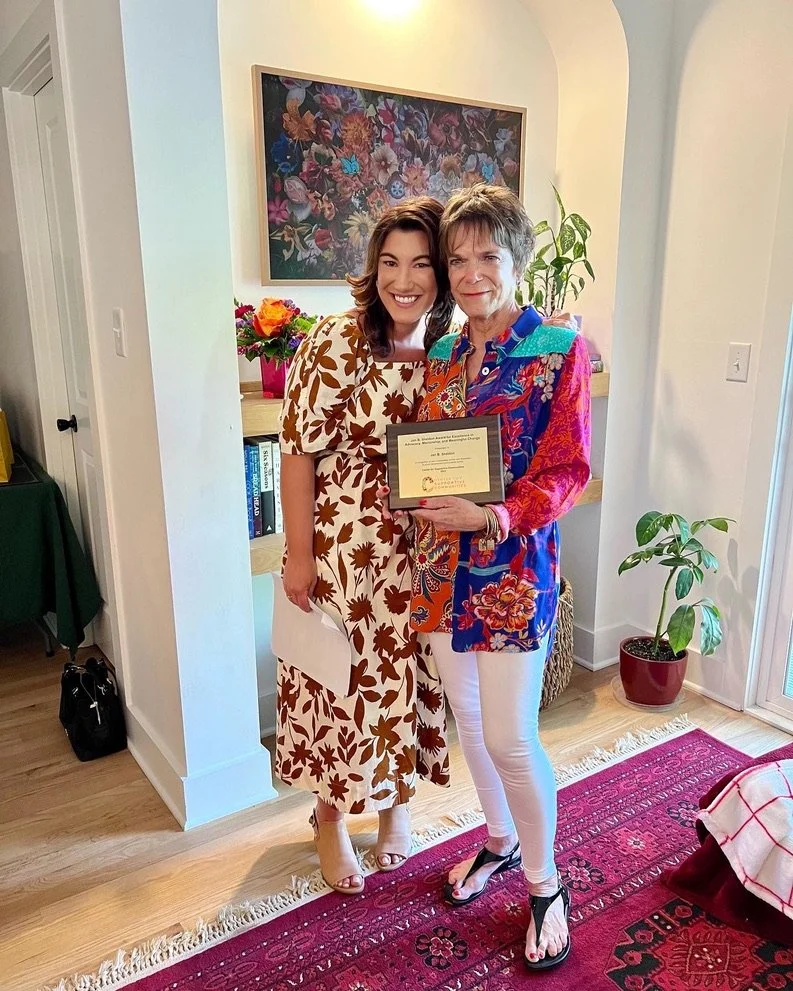 Two women standing together inside a home, one holding a framed certificate. The woman on the left is wearing a white dress with brown floral patterns, and the woman on the right is wearing a colorful floral blazer with white pants and black high hee