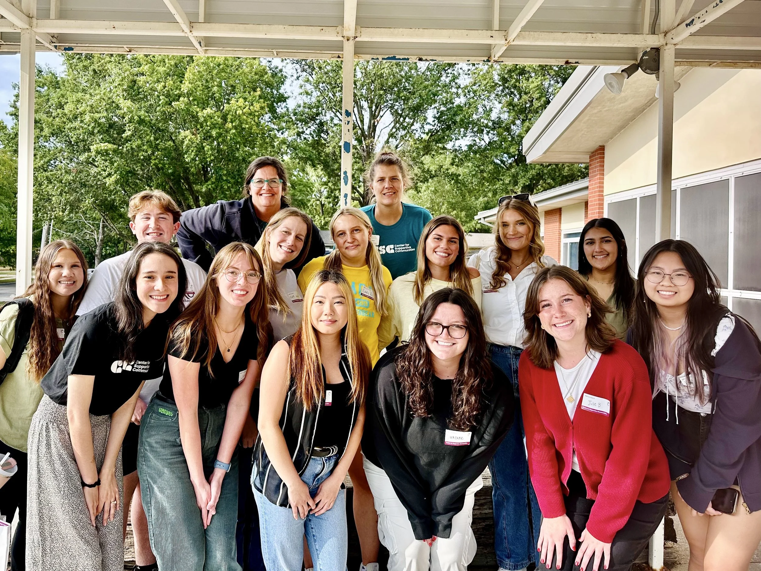 Group of young women and adult women smiling outdoors under a metal awning, with trees and parts of a building in the background.