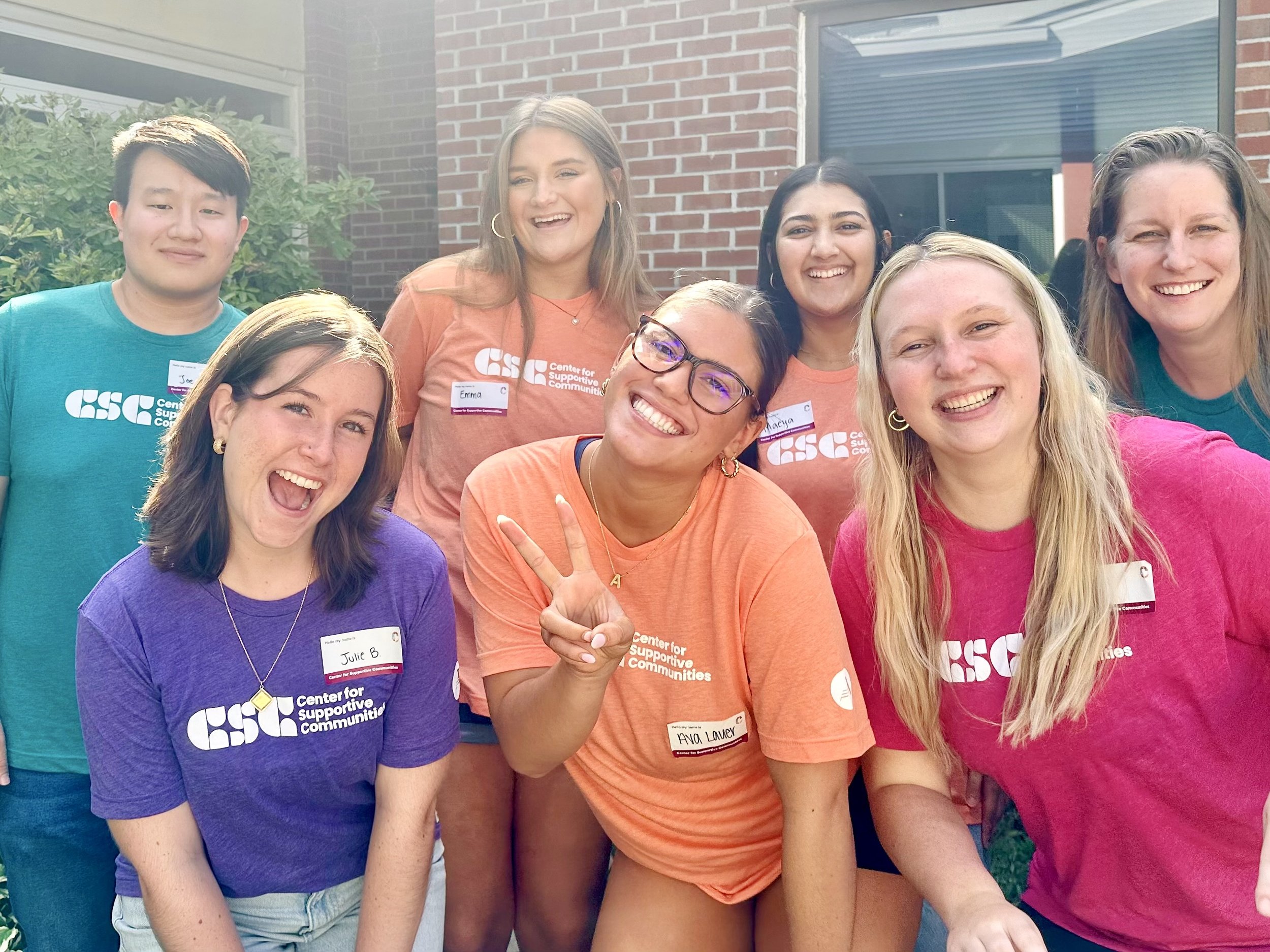 Group of seven diverse young women smiling and posing outdoors in front of a brick building. They are wearing colorful t-shirts with a 'Center for Supportive Communities' logo and name tags.