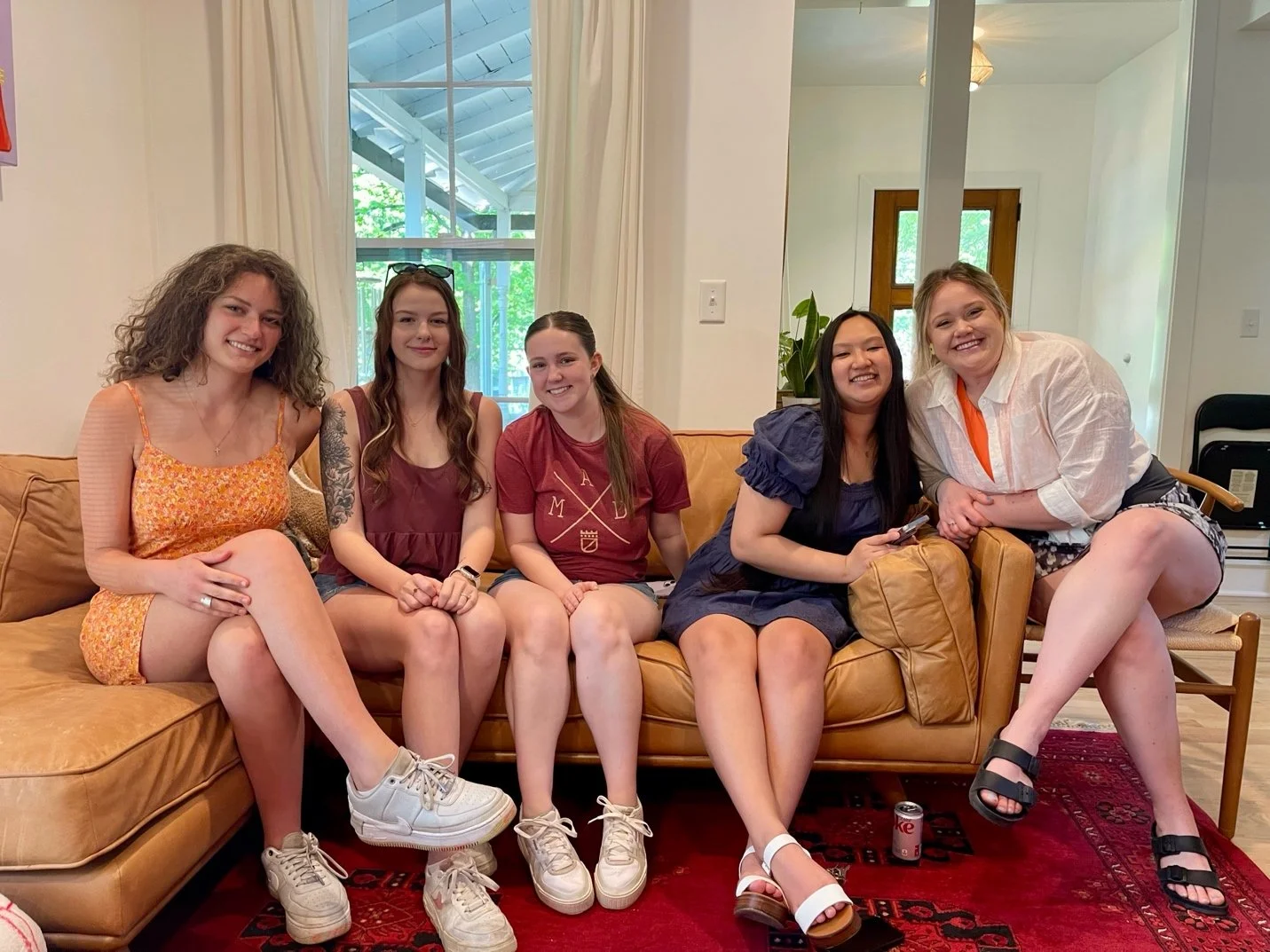 Five young women sitting on a tan couch in a living room, smiling at the camera, with a window behind them and a plant visible through the glass, and a red patterned rug on the floor.