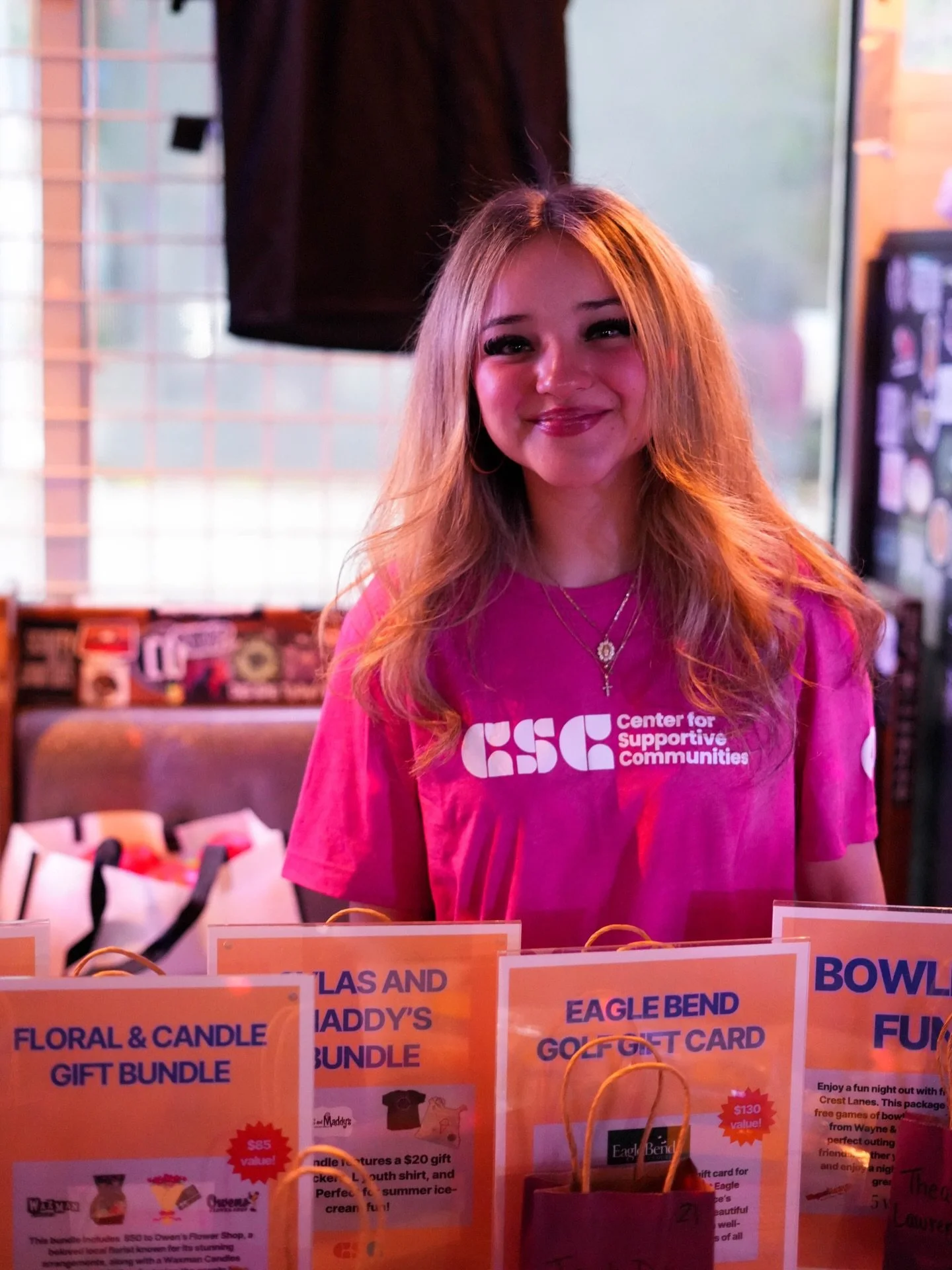 A smiling woman with long red hair wearing a pink t-shirt with a CSC t-shirt volunteering at an event.