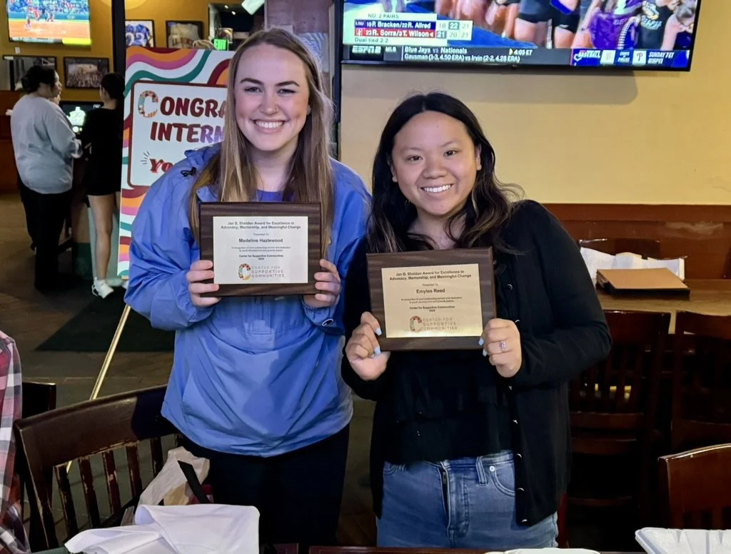 Two young women smiling and holding awards in a restaurant or cafe, with a television screen and a colorful sign in the background.