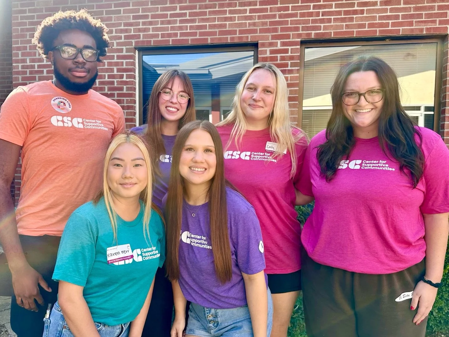 Group of six people standing outside in front of a brick building, wearing colorful t-shirts with the Center for Supportive Communities logo, smiling at the camera.