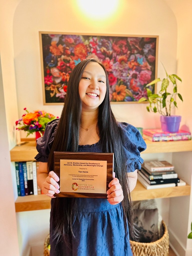 Young woman smiling and holding an award plaque in a room decorated with colorful flowers and books.